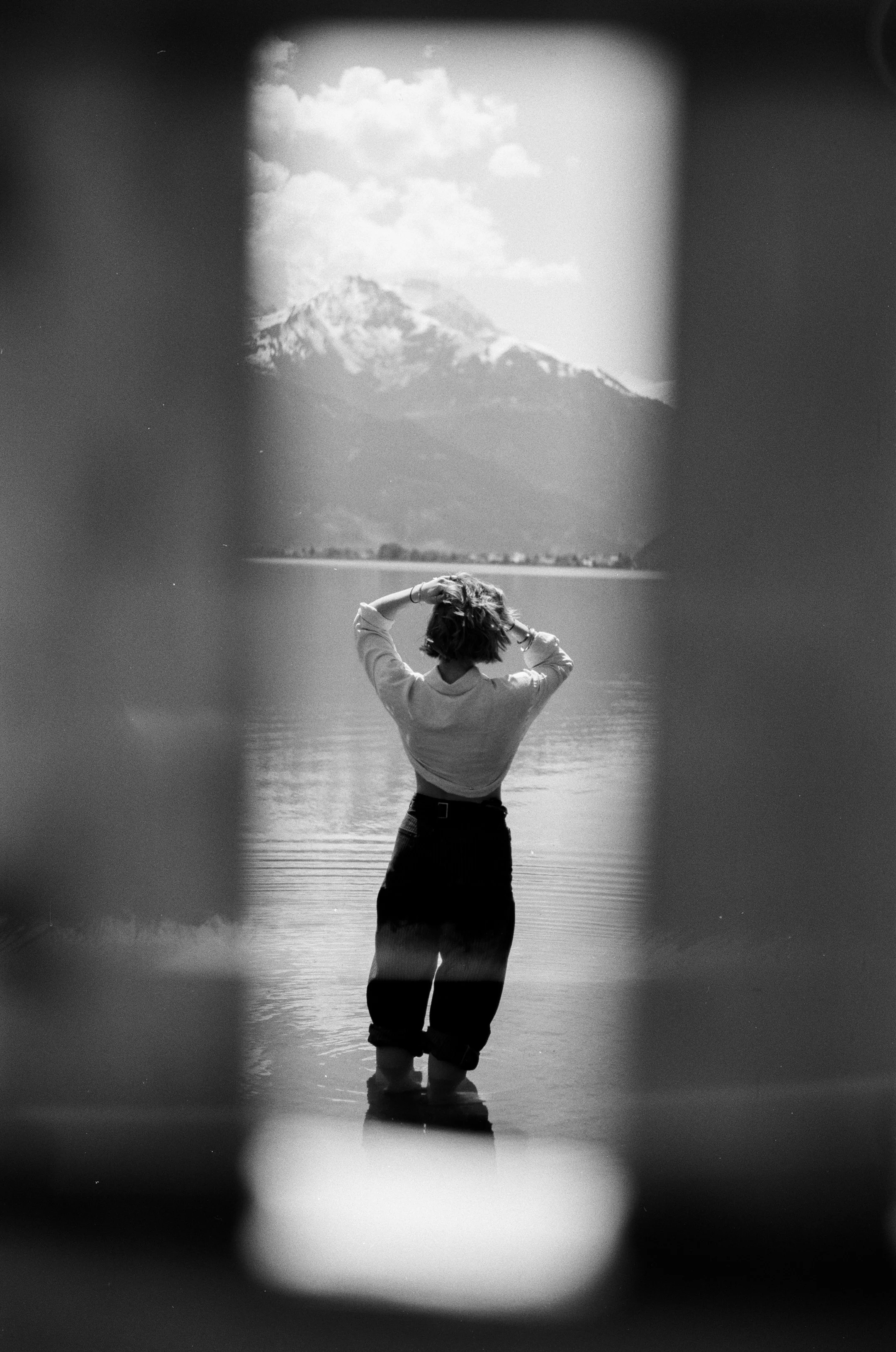Schwarz-weiß-Foto einer Frau, die im Wasser steht, mit Blick auf einen Berg mit Schneegipfel im Hintergrund, durch eine Rahmenöffnung fotografiert.