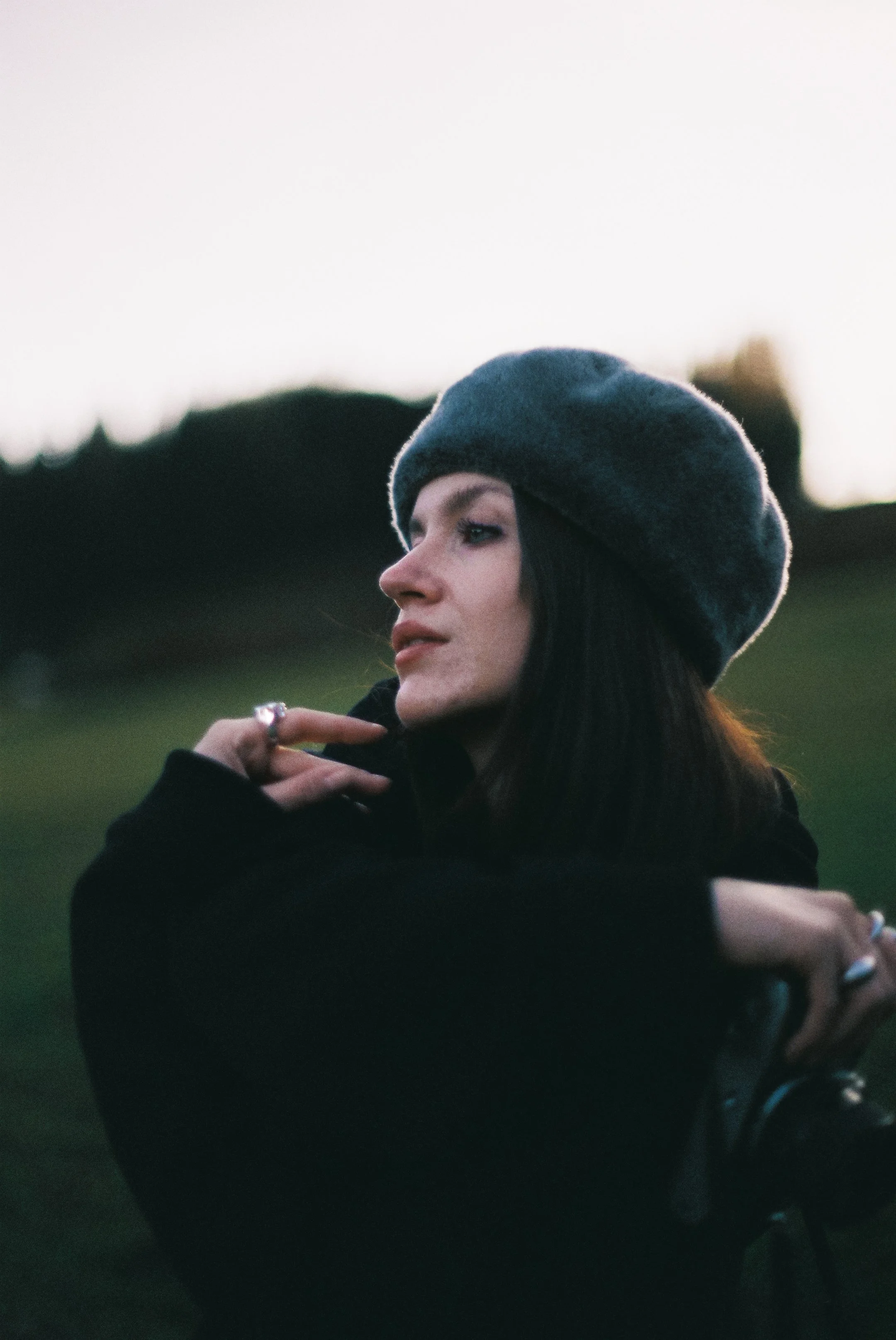 Seitenprofil eines Mädchens mit dunklen Haaren, grauem Hut und schwarzer Kleidung, im Freien bei Dämmerung.