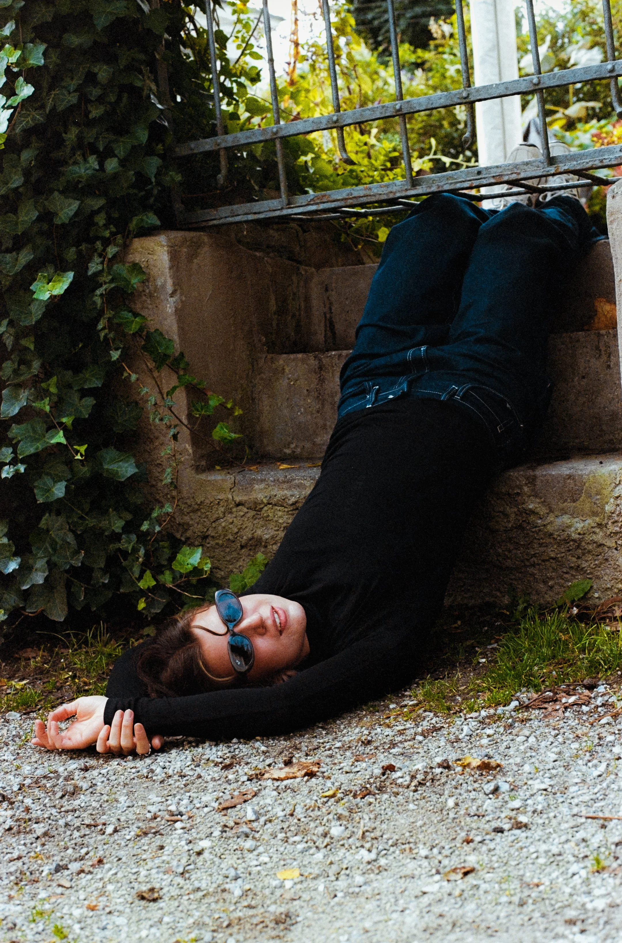 Frau liegt auf dem Boden vor einer Treppe, trägt schwarze Kleidung und Sonnenbrille, umgeben von grünen Pflanzen.