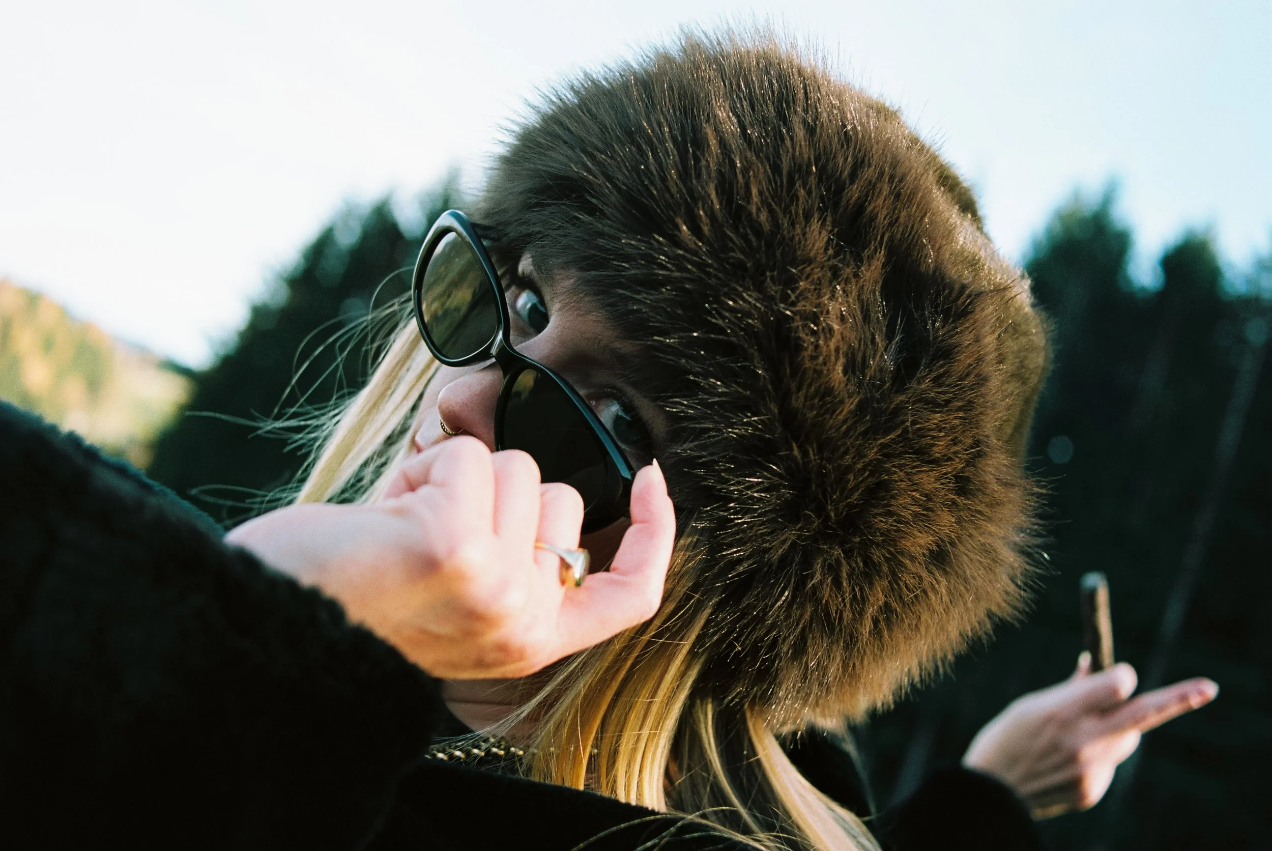 Junge Frau mit dunklen Haaren und Fellmütze, die schwarze Sonnenbrille abnimmt, draußen im Freien, im Hintergrund Bäume und Himmel.