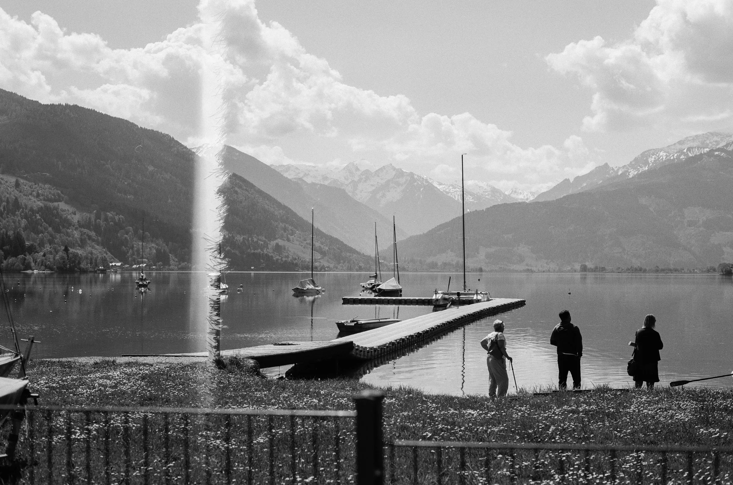 Drei Personen am Ufer eines Sees, mit Bergen im Hintergrund und Segelbooten auf dem Wasser, in Schwarz-Weiß.