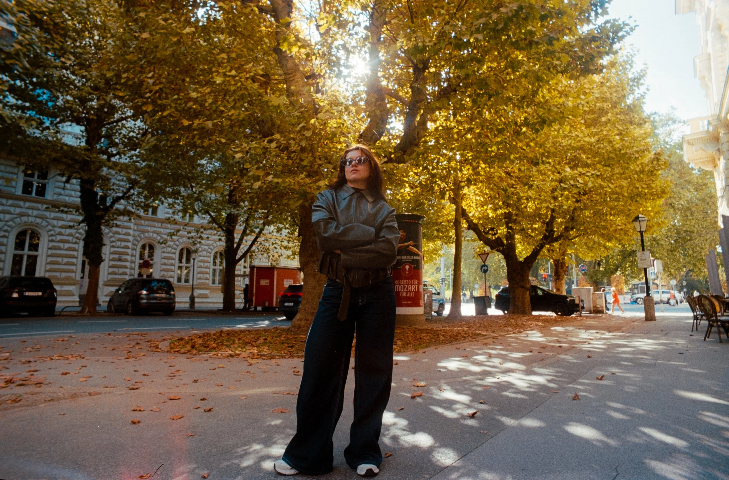 Frau in Sonnenbrille und schwarzer Lederjacke steht mit verschränkten Armen an einem sonnigen Herbsttag in einer Stadt unter goldgelben Bäumen.