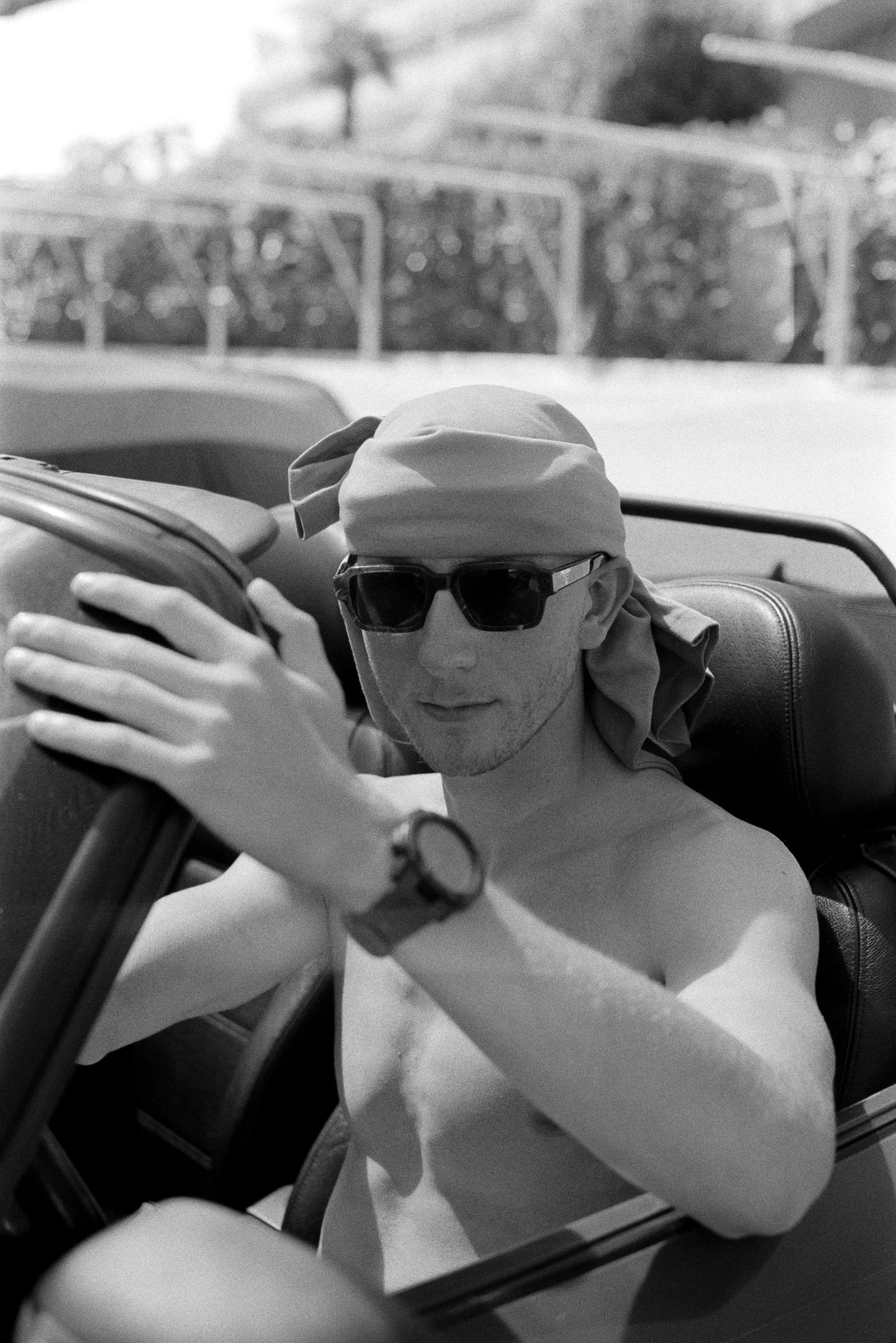 Ein nackter Mann trägt Sonnenbrille, eine Uhr und ein Tuch auf dem Kopf, sitzt in einem offenen Fahrzeug, umgeben von einem Tor im Freien.