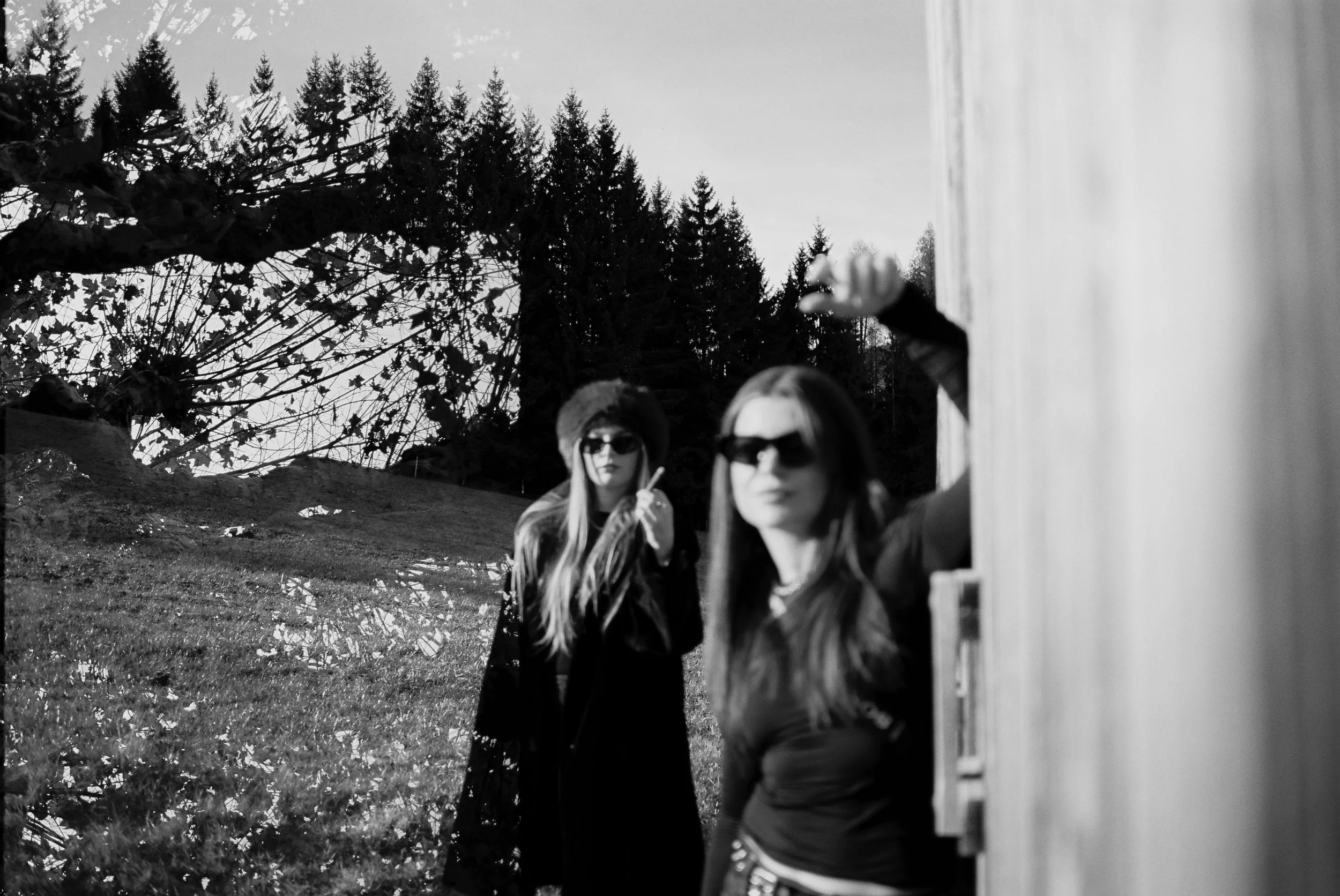 Zwei junge Frauen tragen Sonnenbrillen, eine steht im Vordergrund, die andere im Hintergrund, während sie an einem Haus außerhalb eines Waldes stehen.