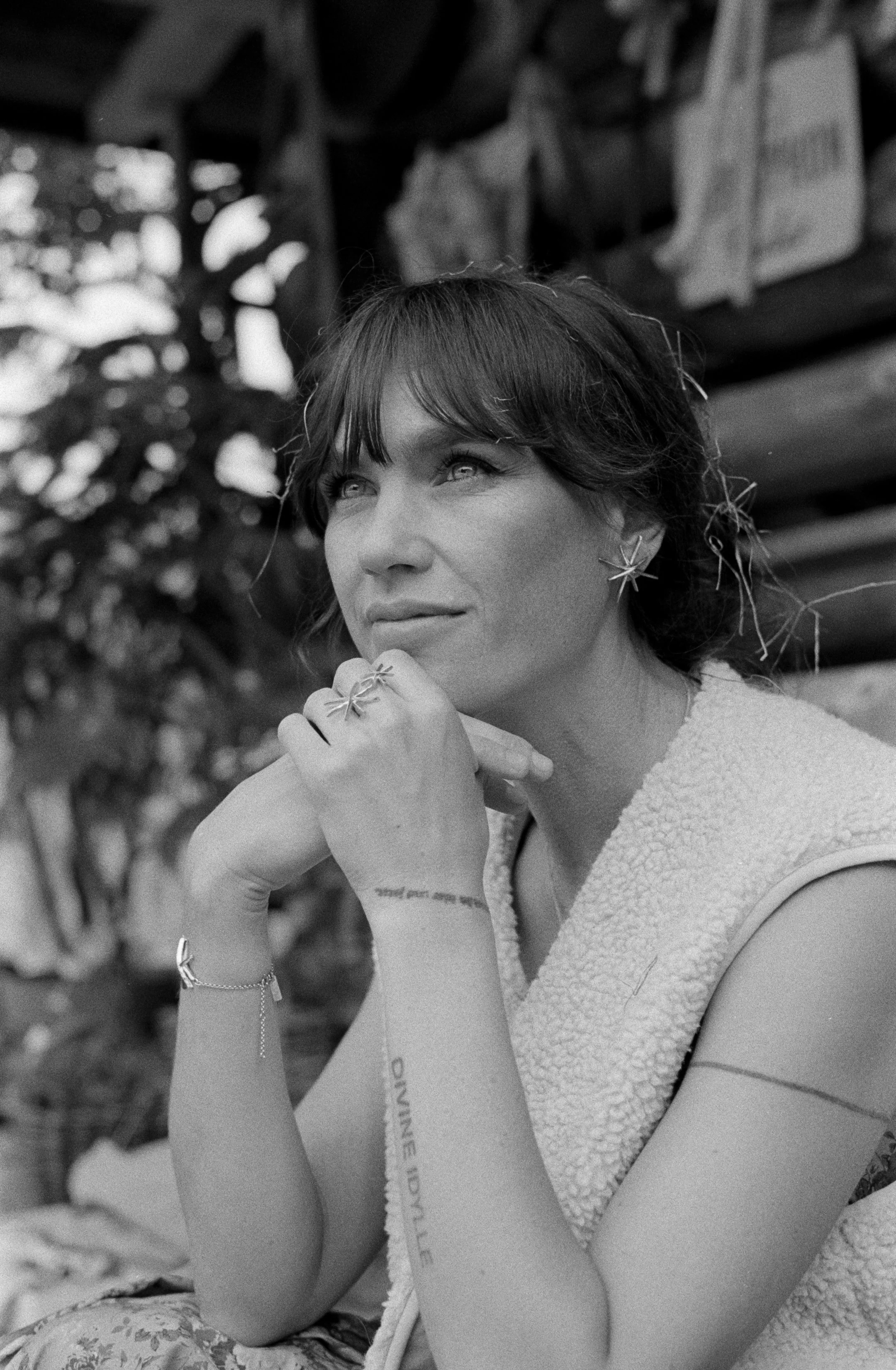 Schwarz-Weiß-Foto einer Frau mit kurzen Haaren, die nachdenklich aussieht. Sie trägt Blumenringe, Armbänder und hat Tattoos am Arm. Im Hintergrund sind Bäume und ein Holzregal zu sehen.