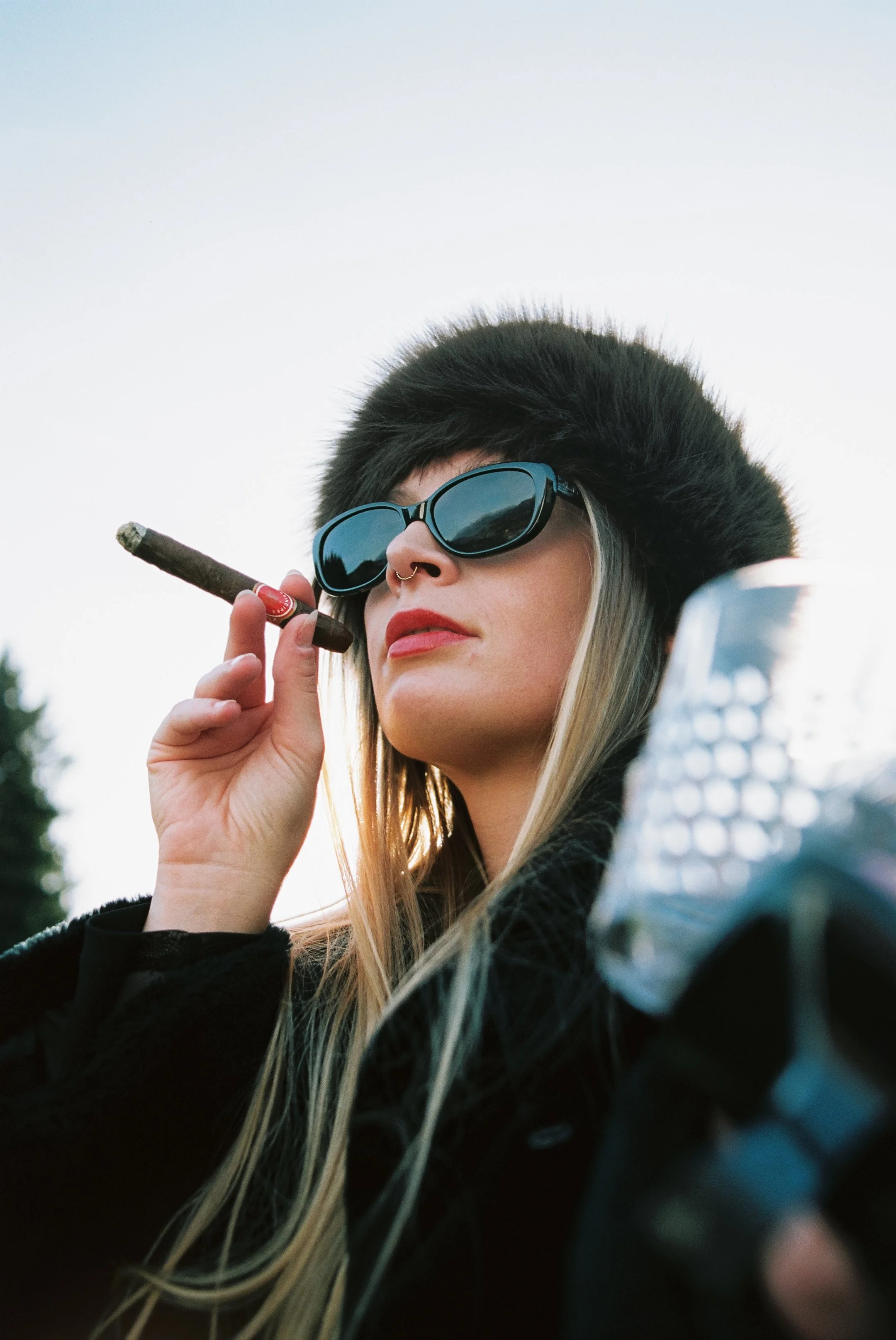 Eine Frau trägt eine schwarze Pelzmütze, dunkle Sonnenbrille und raucht eine Zigarre im Freien.