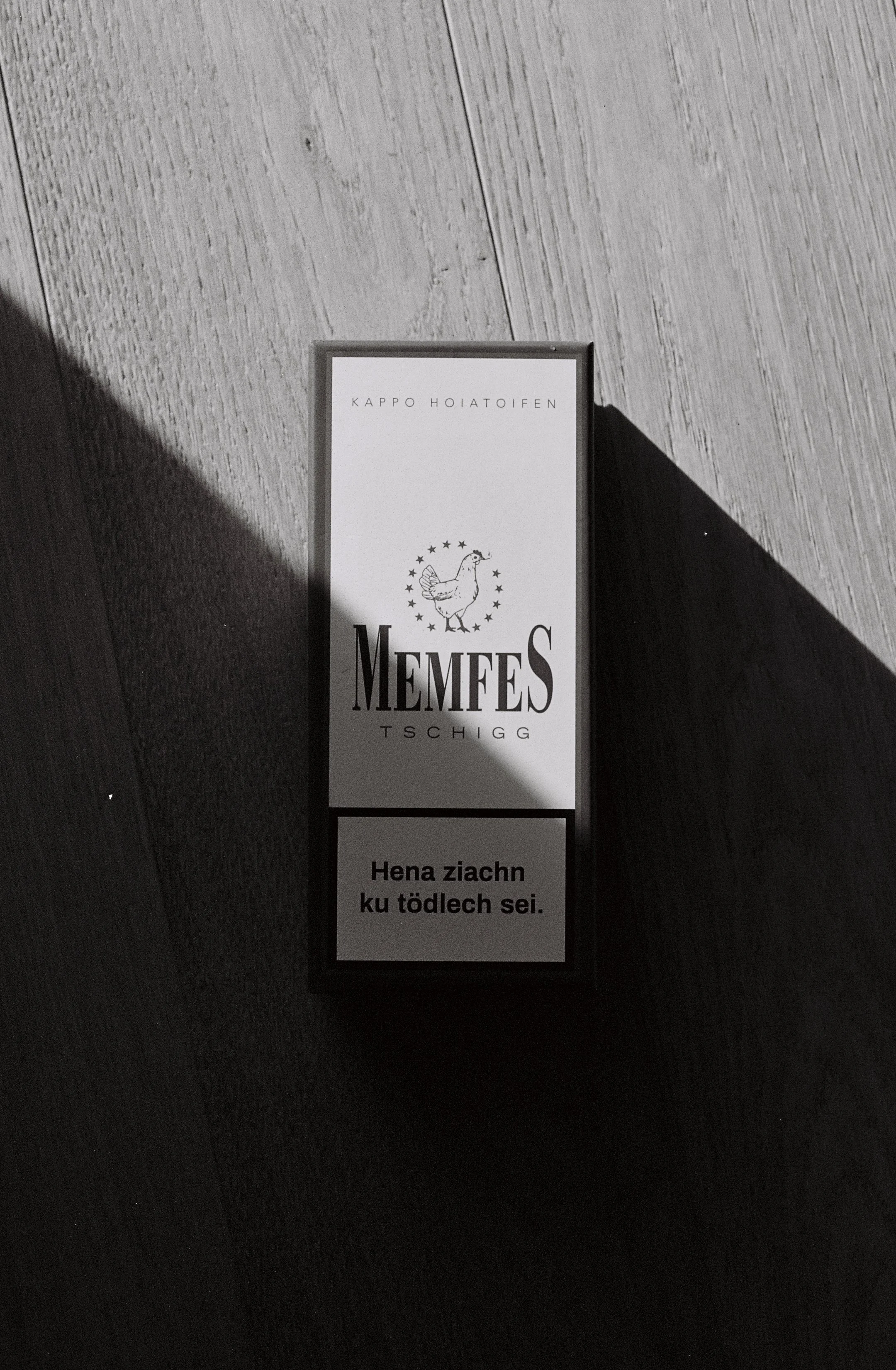 Schachtel Zigaretten mit Aufschrift 'MEMEES' und einem Hahn, auf einem Holzboden liegend, schwarz-weiß fotografiert.