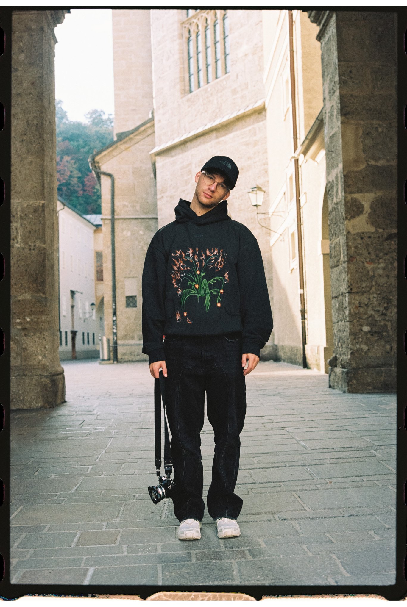 Ein junger Mann steht auf einer Kopfsteinpflasterstraße vor alten Stein- und Backstein-Gebäuden. Er trägt eine schwarze Kapuzenjacke mit einer bunten Blumen- und Feuer-Design, schwarze Hosen, weiße Sneakers, eine schwarze Cap und hält eine Kamera in der Hand.