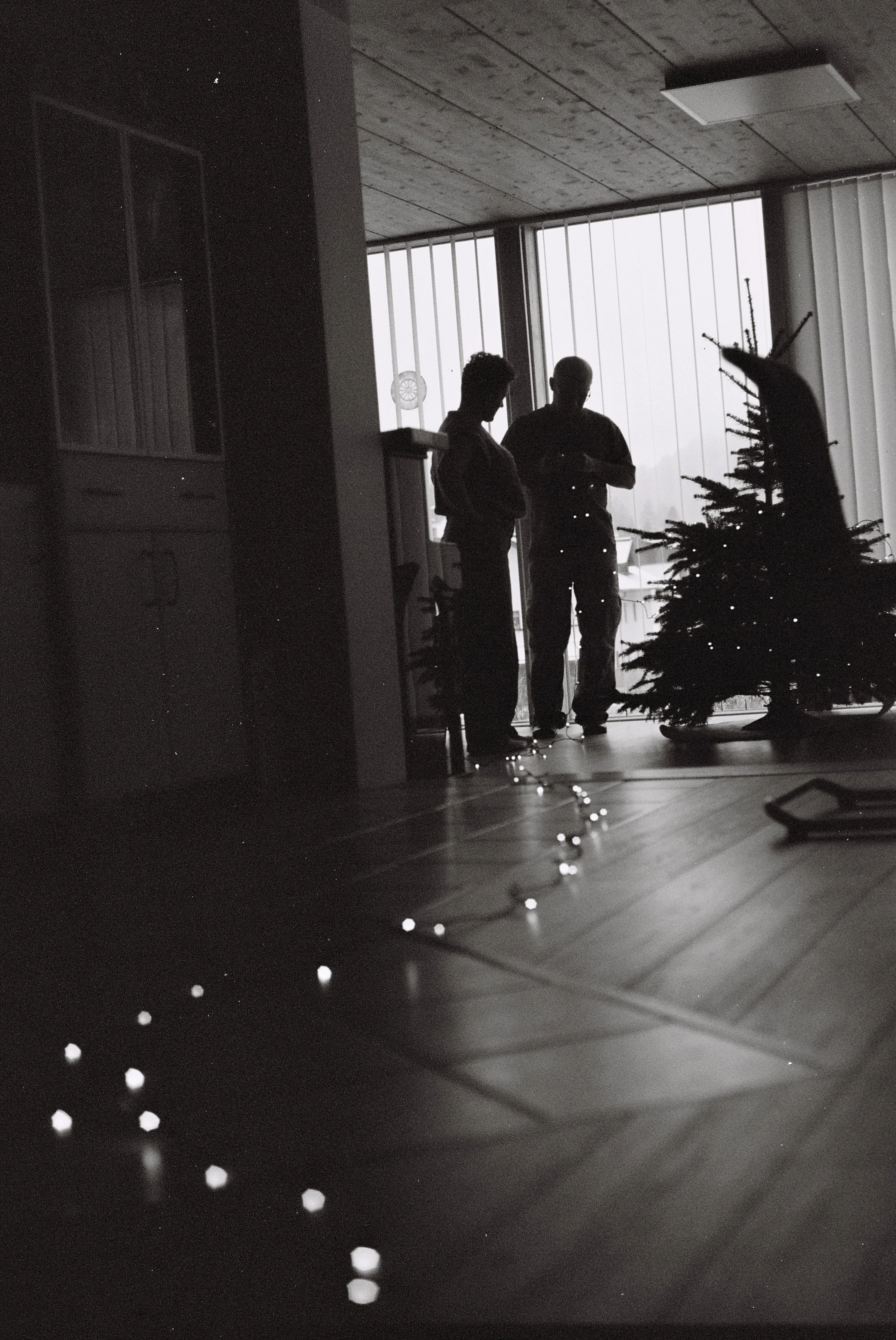 Zwei Menschen im Silhouettenstil beim Weihnachtsbaum dekorieren in einem Raum mit großem Fenster.