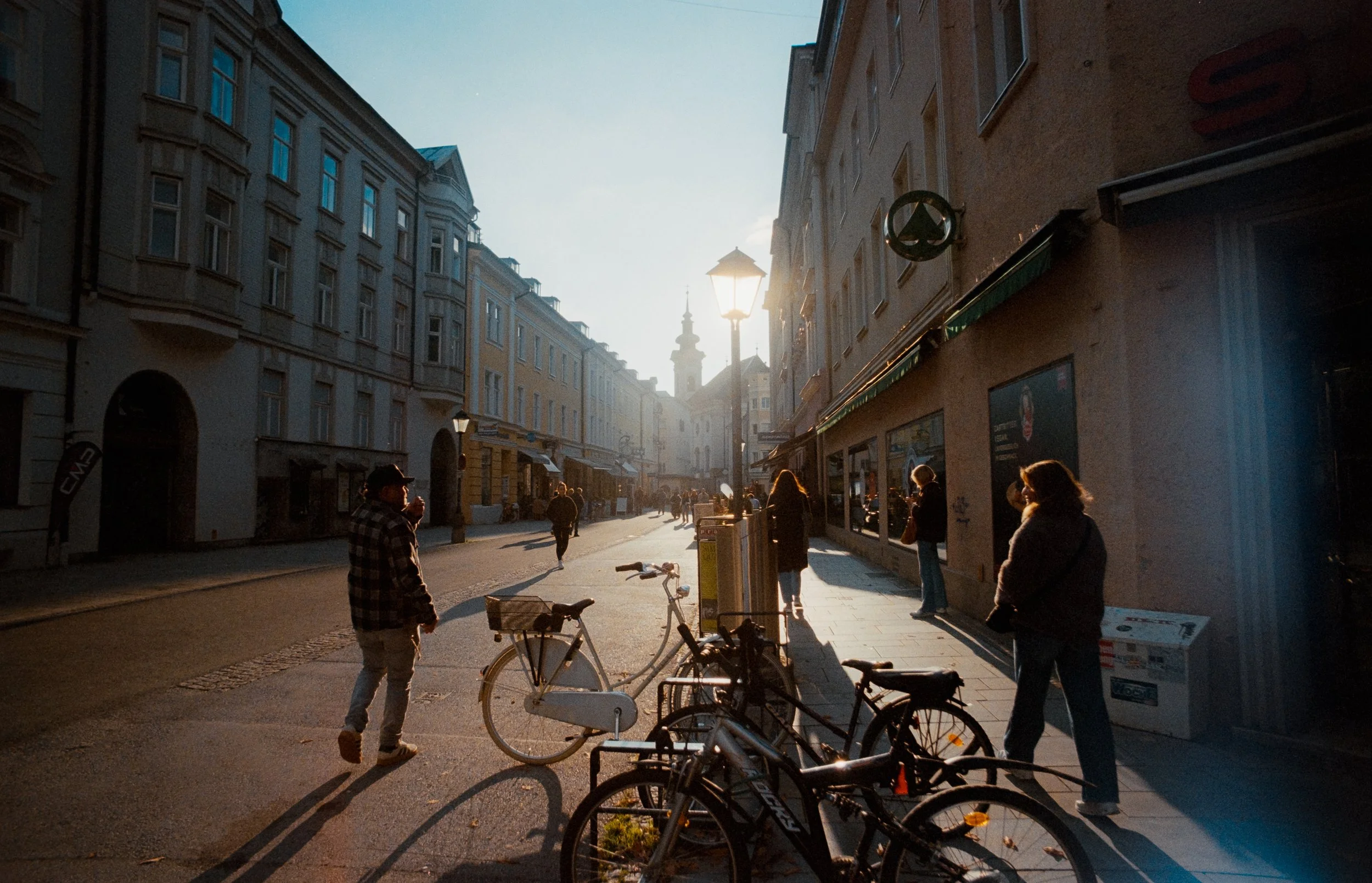 Menschen gehen an einer Straßenecke in einer Stadt im Sonnenuntergang vorbei, Fahrräder parken an der Seite, leuchtender Himmel mit einem Turm im Hintergrund.