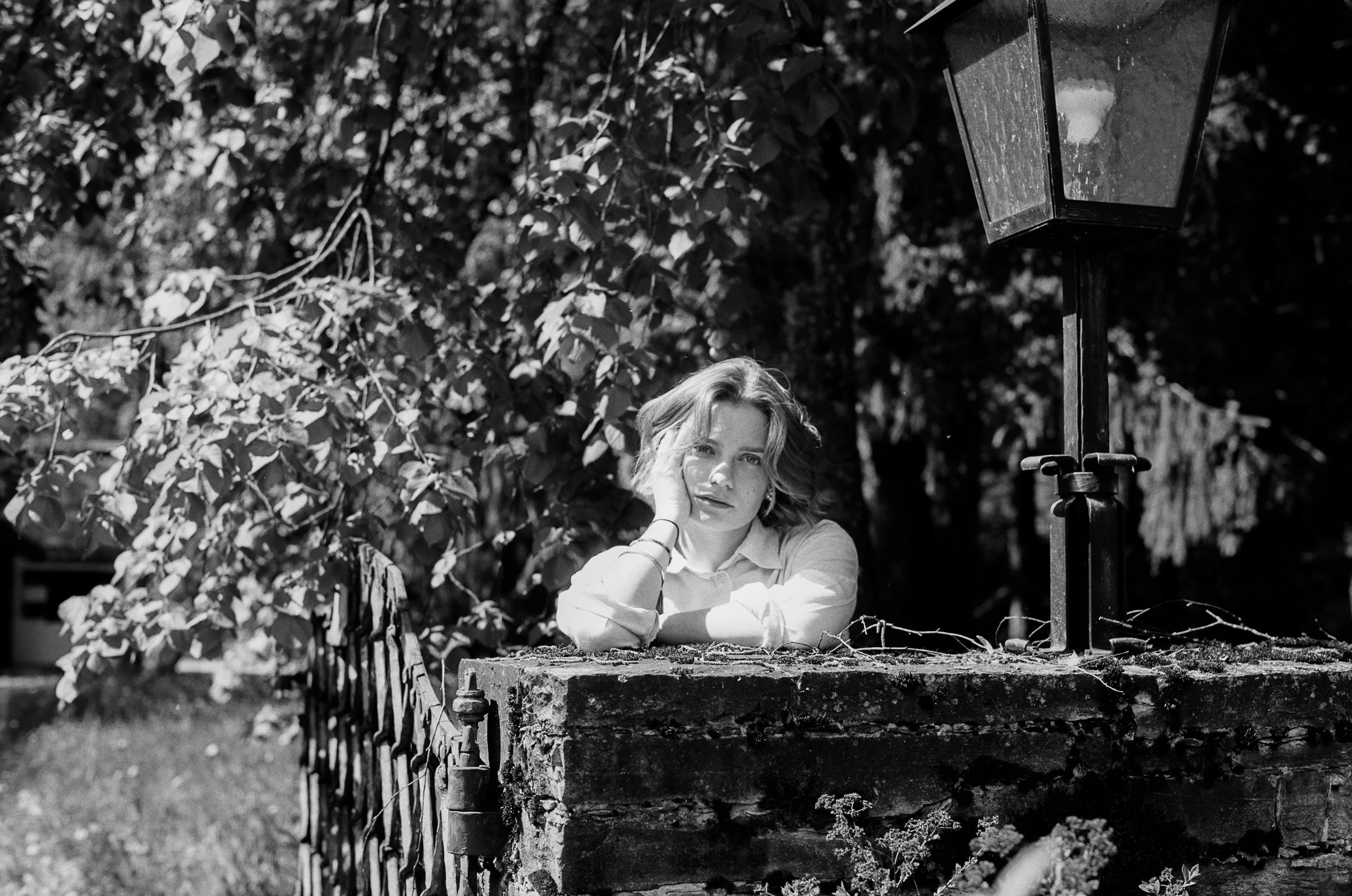 Schwarz-weiß-Foto eines jungen Mädchens, das an einer bröckelnden Steinmauer lehnt, umgeben von Bäumen, bei Sonnenlicht, mit einem Laternenmast im Hintergrund.