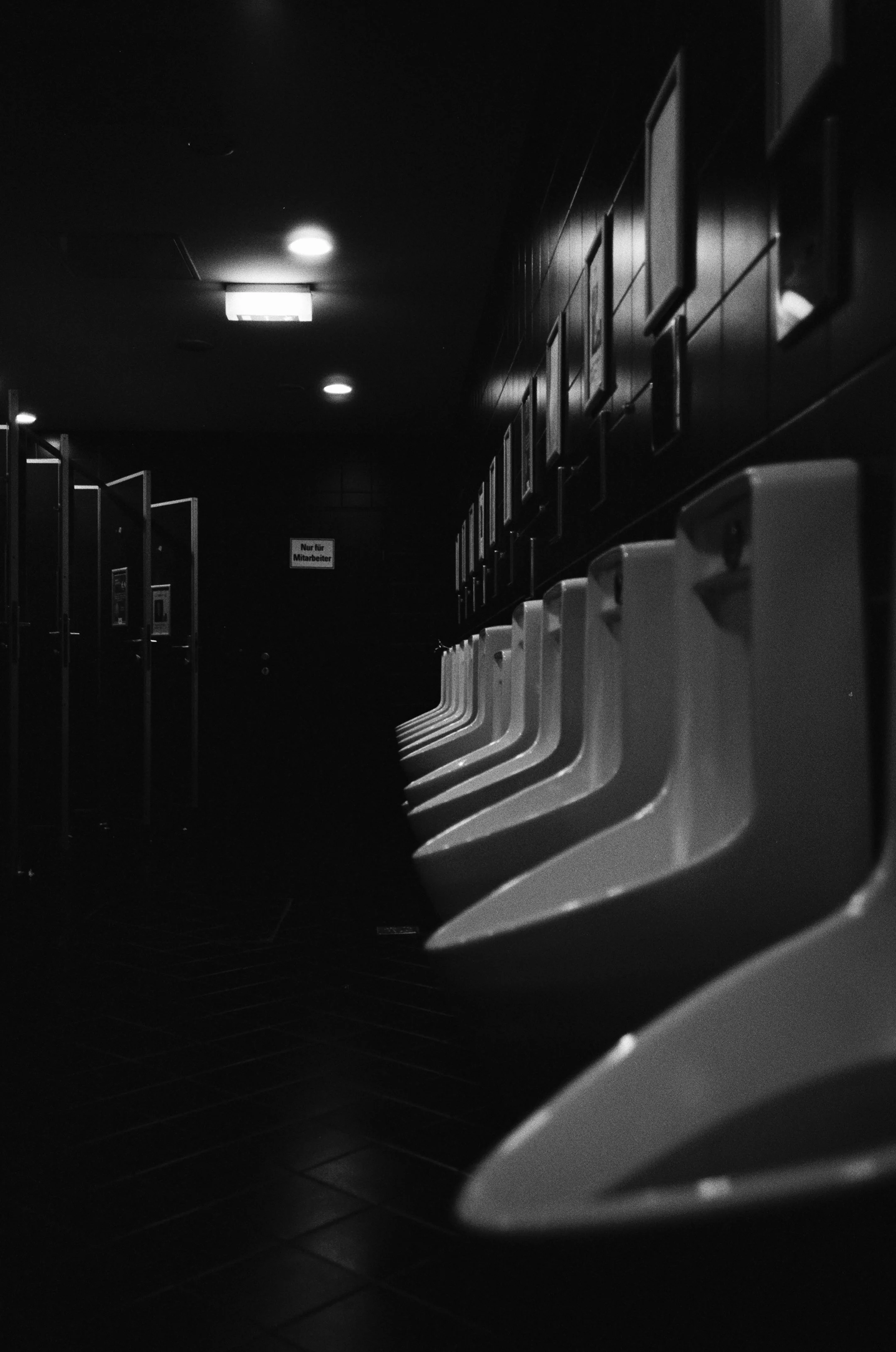 Ein dunkler Eingangsbereich mit einer Reihe von Urinalen auf der rechten Seite und einer Tür im Hintergrund, die mit einem kleinen rechteckigen Licht überstrahlt ist.