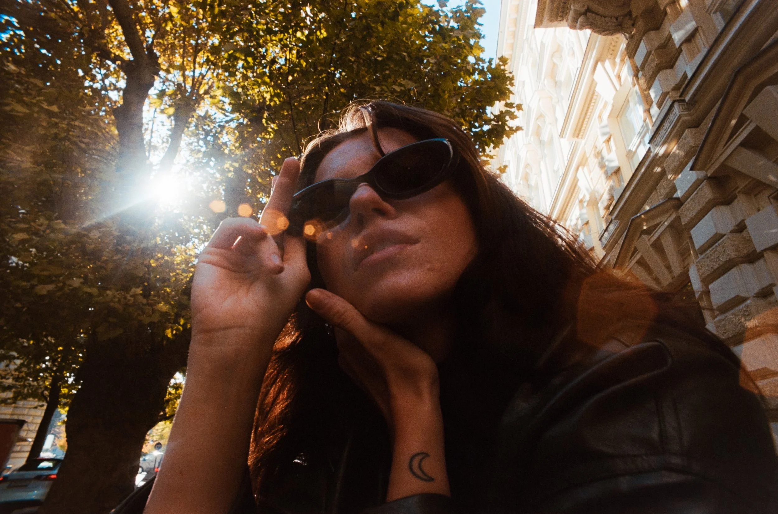Junge Frau mit schwarzen Sonnenbrillen, Sonnenlicht hinter Bäumen, hinter Gebäude in einer Stadt