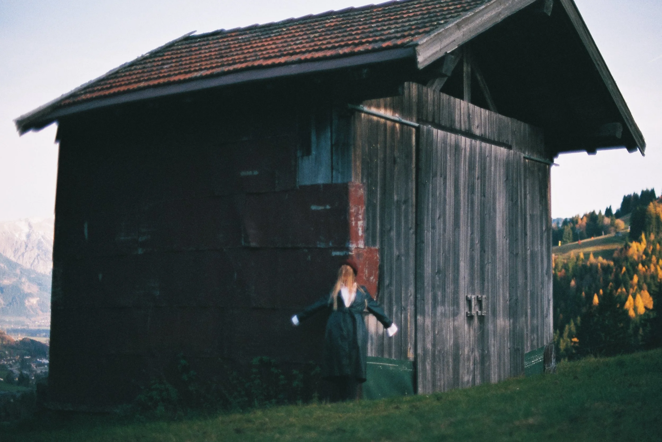 Ein altes Holzschuppen auf einer Hügelwiese mit einem modellhaften weißen Mädchen davor, im Hintergrund Berge und Bäume mit herbstlicher Laubfärbung.
