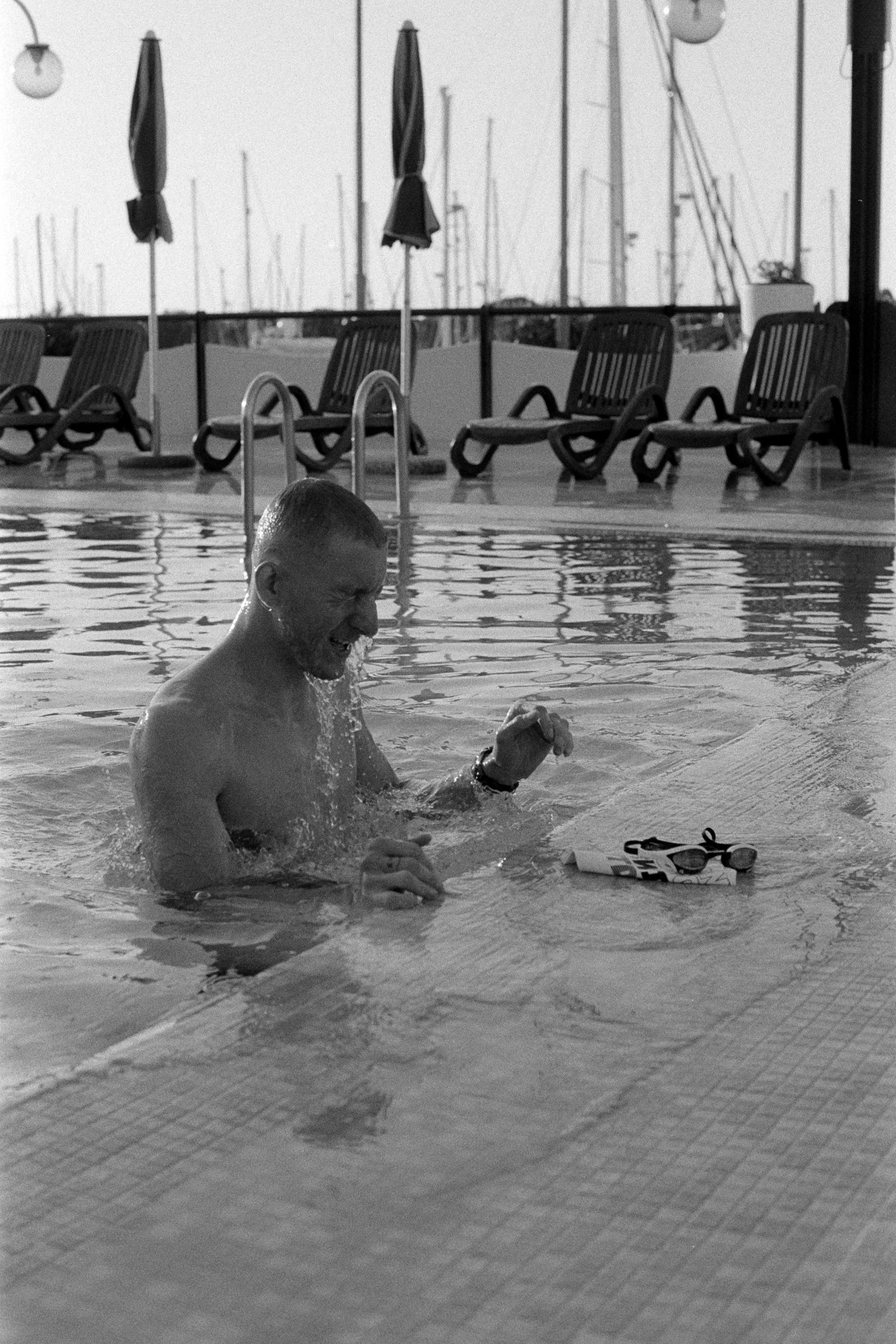 Ein Mann im Schwimmbad lacht und freut sich, während er eine Wasserflasche mit Schwimmbrille auf dem Rand des Pools ablässt. Im Hintergrund sind Liegestühle und Boote im Hafen sichtbar.