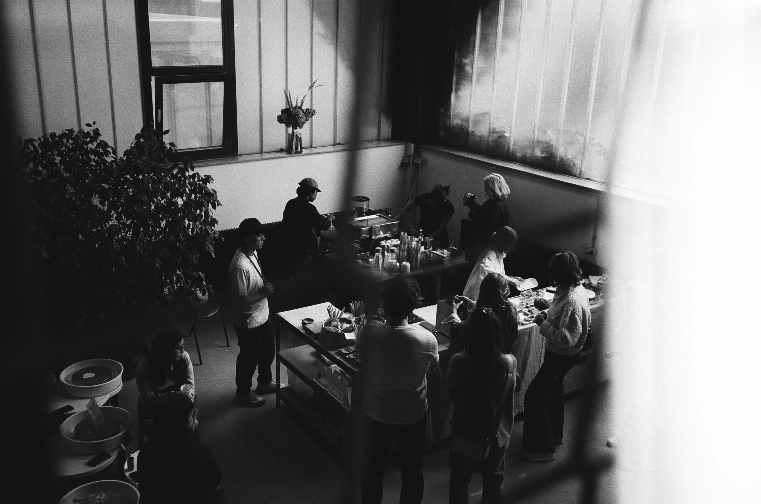 Menschen in einem Raum beim Essen und Fotografieren, schwarz-weiß, mit Pflanzen und großen Fenstern im Hintergrund.