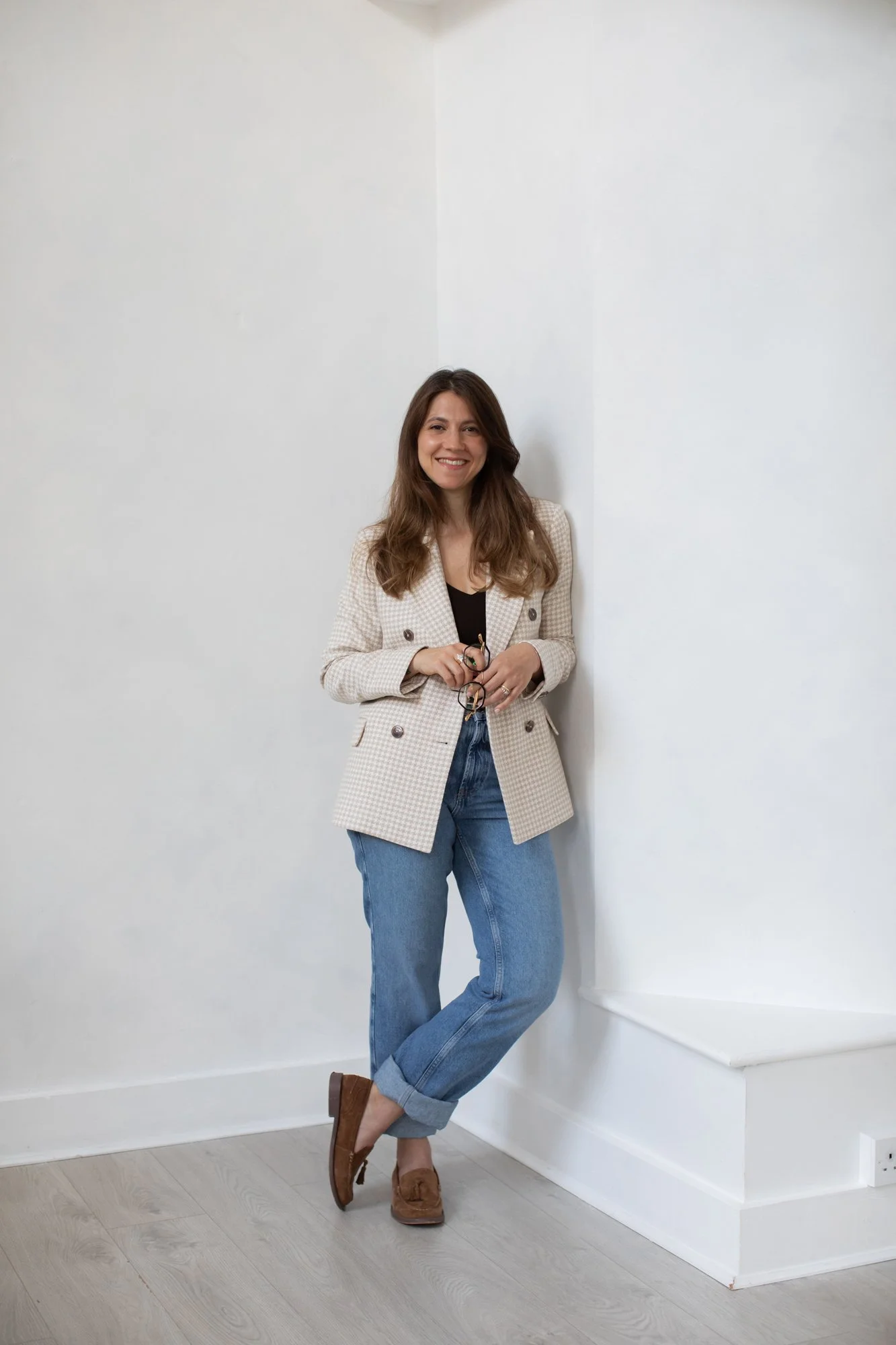 Nergis leaning on a wall, holding her glasses and smiling at the camera