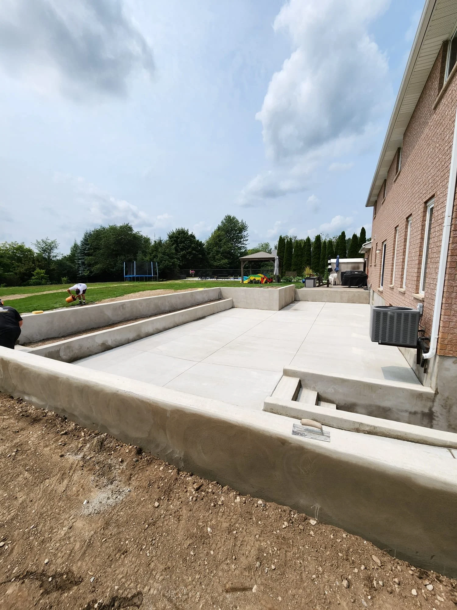 Newly constructed concrete patio area with retaining walls next to a brick house, showing a backyard with grass, trees, and outdoor play equipment under a partly cloudy sky.
