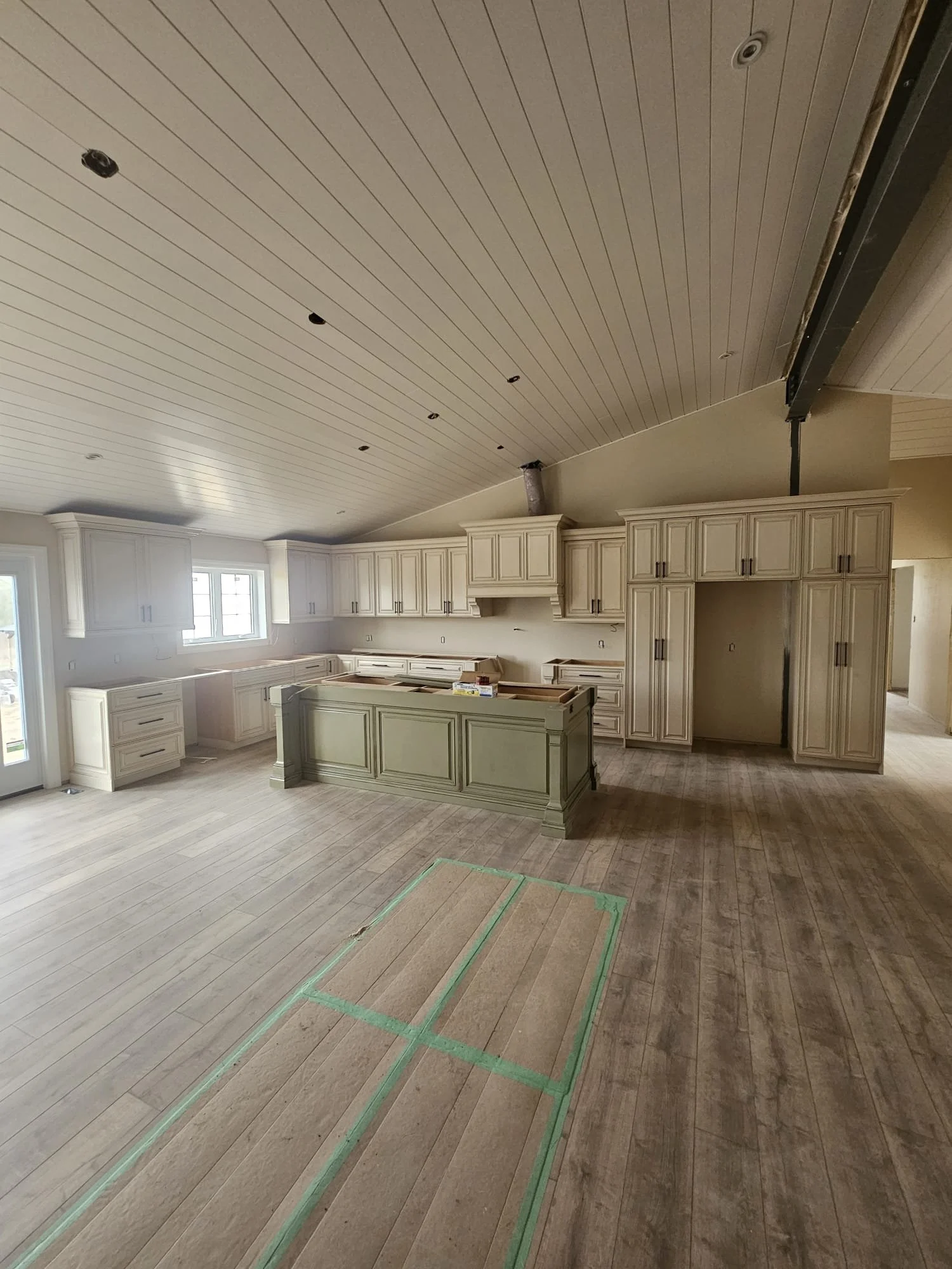 Kitchen under construction with white cabinets, a central island, and unfinished ceiling fixtures. Wooden flooring and windowed doors are visible.