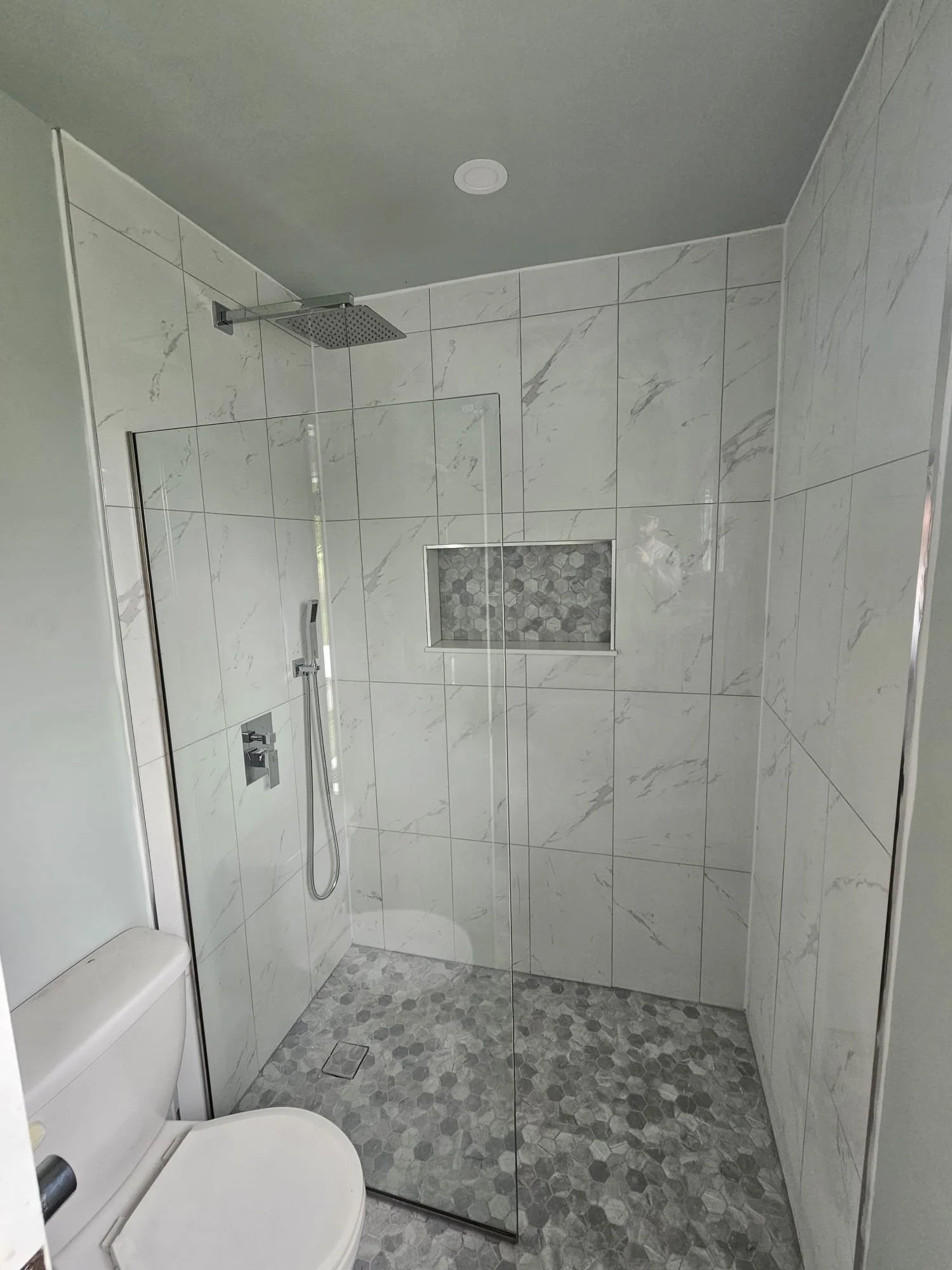 Modern walk-in shower with marble tiles, a rain showerhead, a handheld shower, and a built-in niche with hexagonal tiles, separated from the toilet by a glass partition.