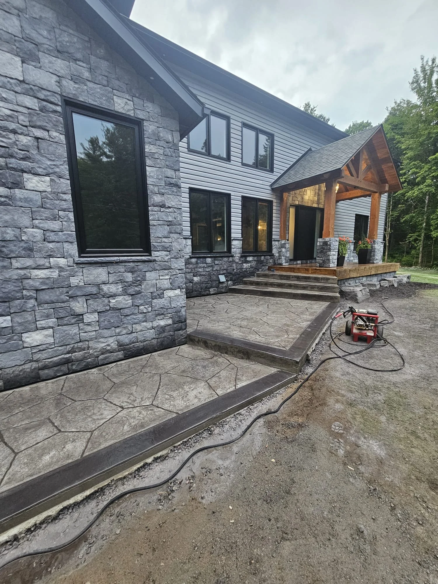 Newly constructed house with stone and metal siding, a covered front porch with wooden beams, steps leading up to the porch, and a concrete patio area. A power washer is on the ground near the patio.