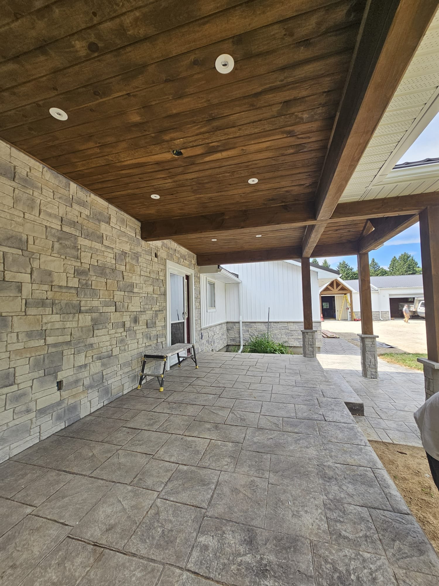 Covered patio with stone flooring, stone and white siding walls, wooden ceiling with recessed lighting, and wooden columns.