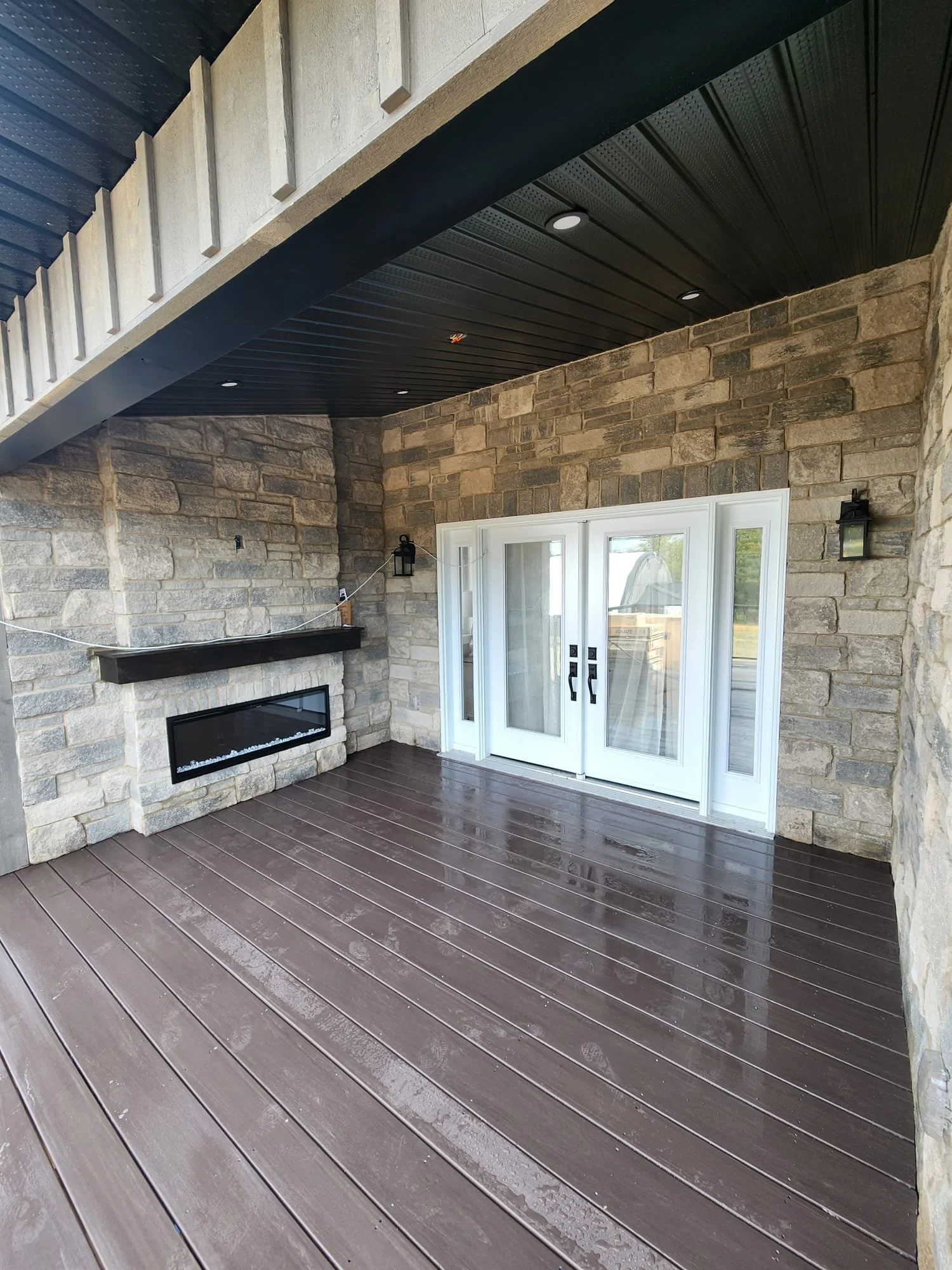 Covered patio area with a wet, dark brown wooden floor, stone walls, a fireplace with a black mantle, white French doors, and black outdoor wall lanterns.