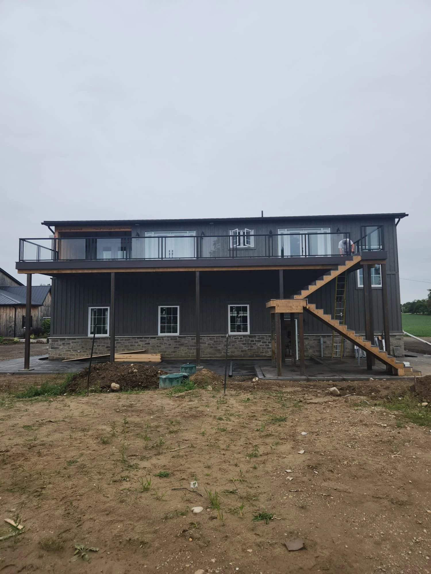 A two-story house under construction with a black exterior, stone foundation, and a large balcony on the upper floor. Construction materials and tools are visible around the site.