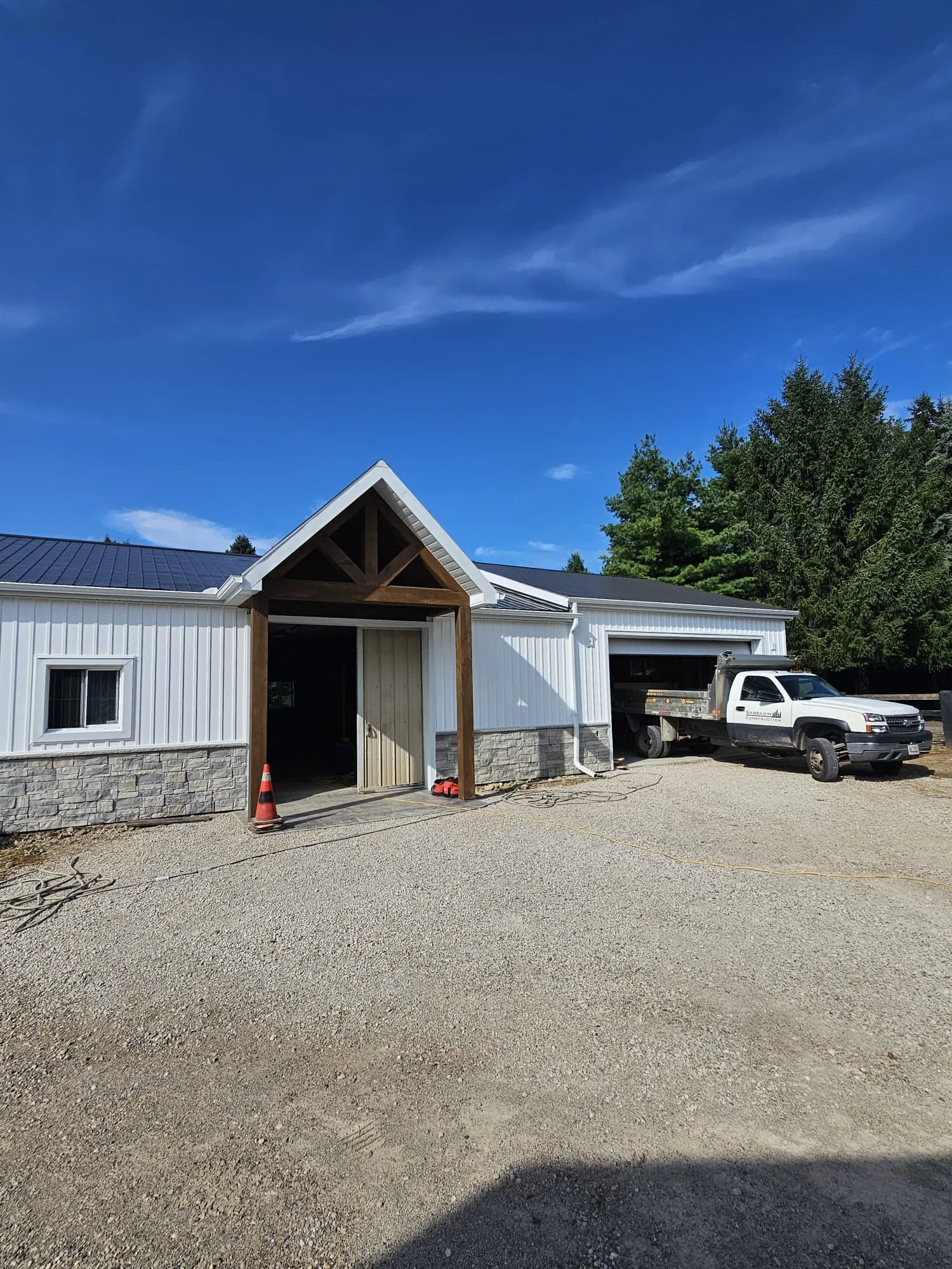A building under construction with a white exterior, stone foundation, and a steep gable roof. A truck is parked in front near a garage door, and construction cones are visible near the entrance.