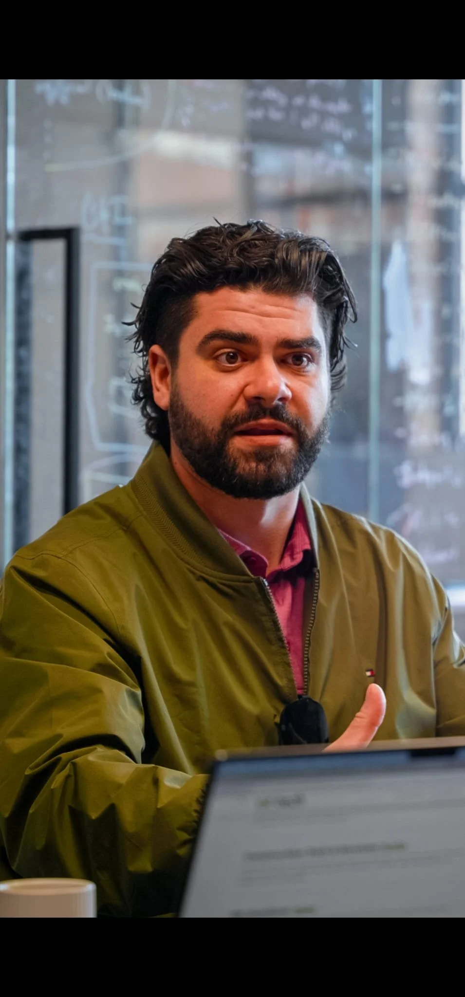 A man with dark hair and a beard sitting at a table in a cafe or restaurant, wearing a green jacket over a maroon shirt, speaking while gesturing with his thumb up. In the foreground, there is a laptop and a coffee cup.