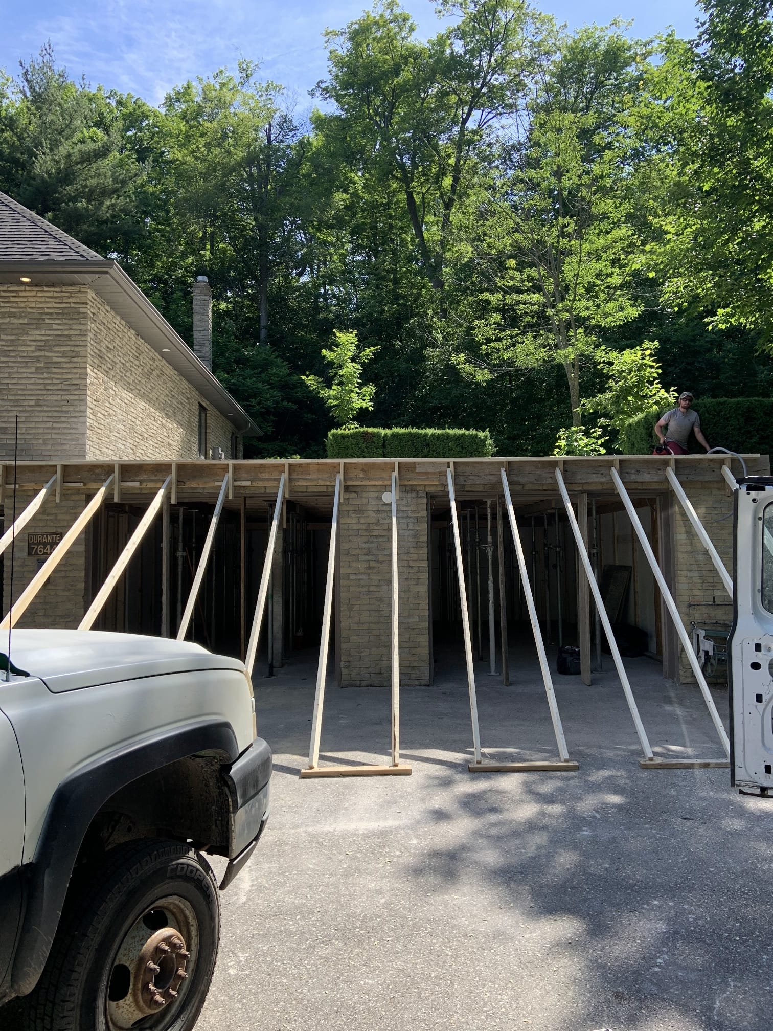 Construction site showing an unfinished building with wooden framing, a worker on top, and a pickup truck parked in the foreground. The background features green trees and a brick house.