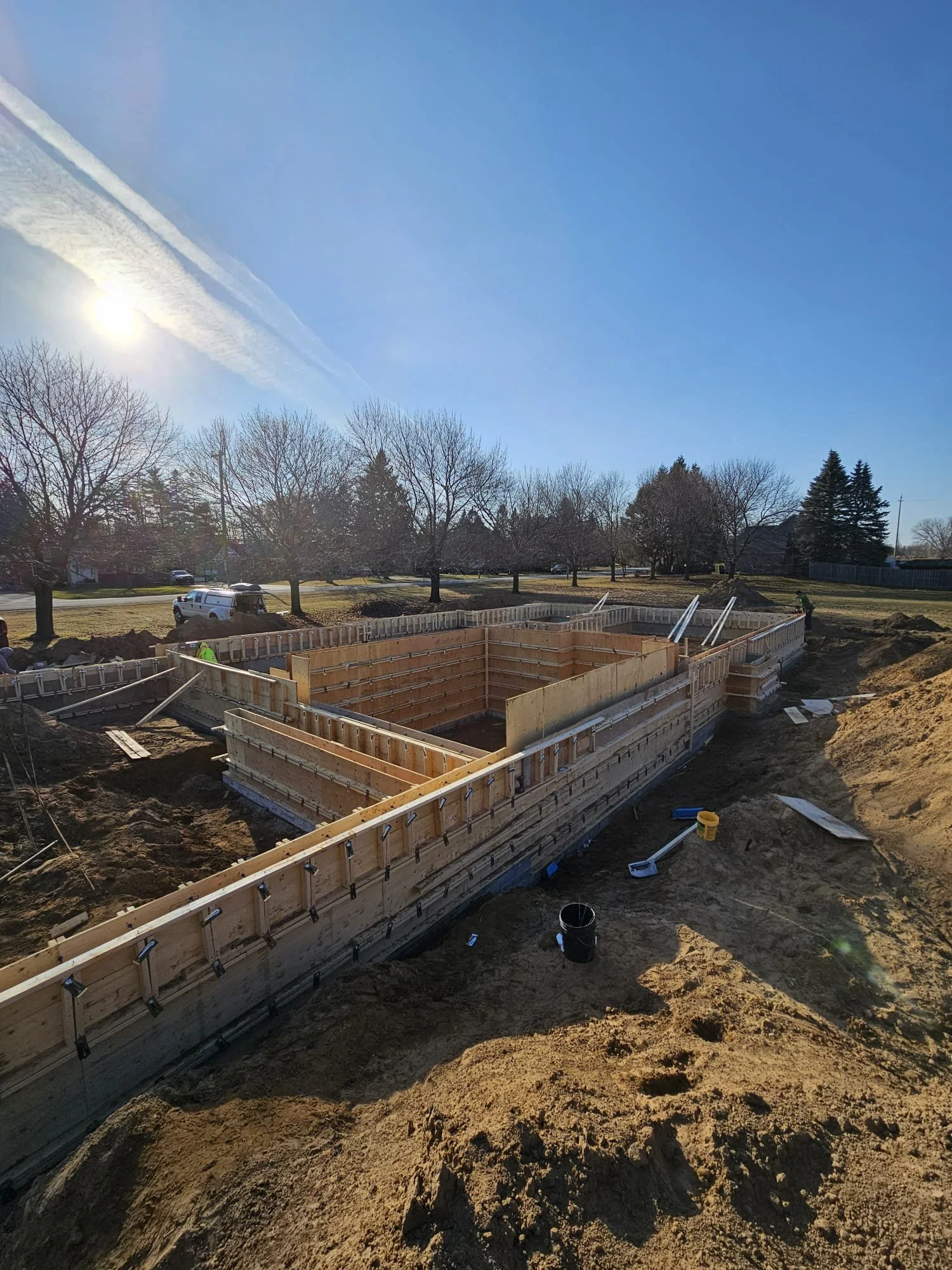 Construction site with wooden framework for building foundation, construction workers, tools, and dirt mounds, under a clear blue sky with the sun.