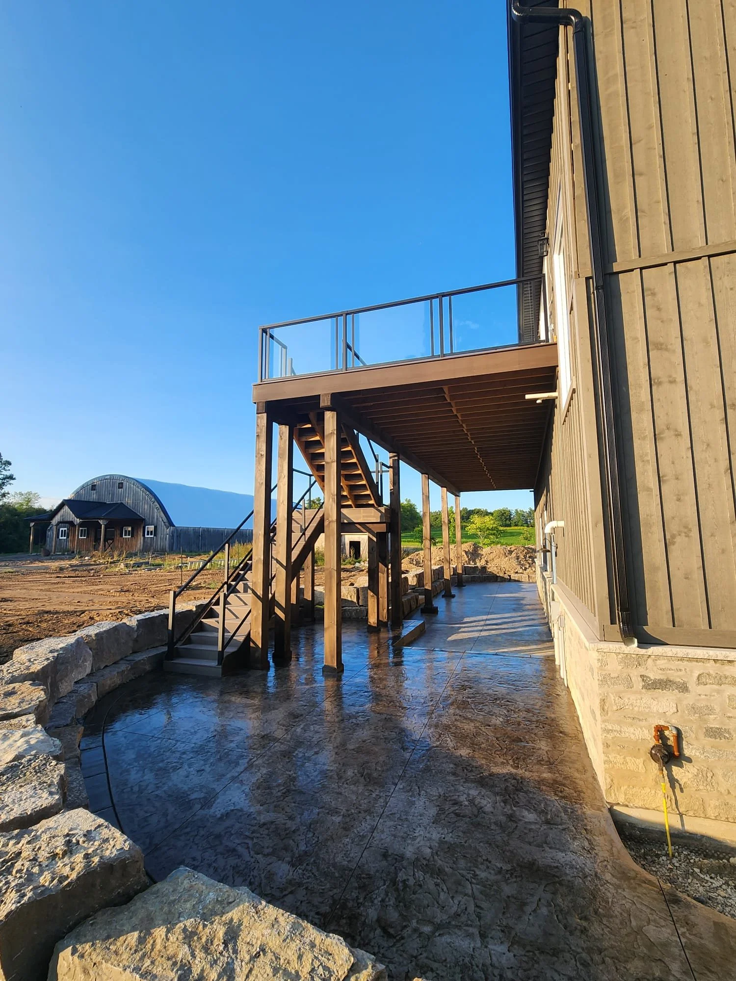 Backyard area with a concrete patio, wooden stairs leading to a second-story balcony, and a black metal railing, with a large wooden building and a barn in the background under a clear blue sky.