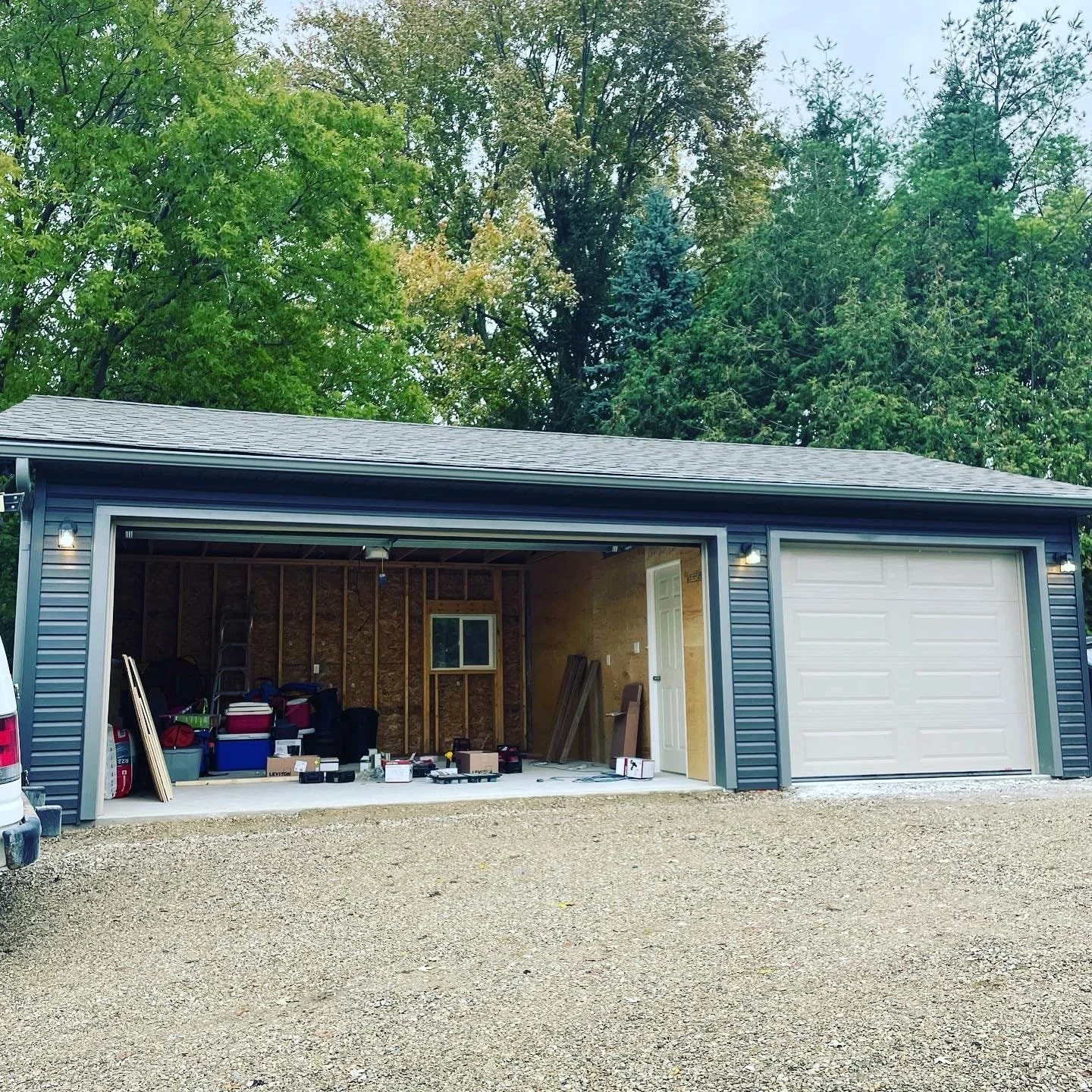 Image shows a garage under construction with tools and supplies inside. The garage has blue siding, a gray roof, and a gravel driveway in front.