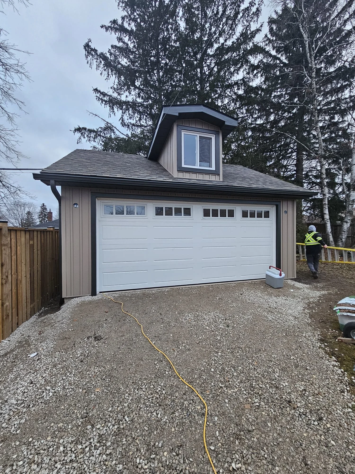 A newly constructed detached garage with a white roll-up door, small windows at the top, beige siding, and a gabled roof. There is a worker on the right side wearing a reflective vest, black jacket, white helmet, and dark pants. The ground in front of the garage is gravel, and a house with trees can be seen in the background.