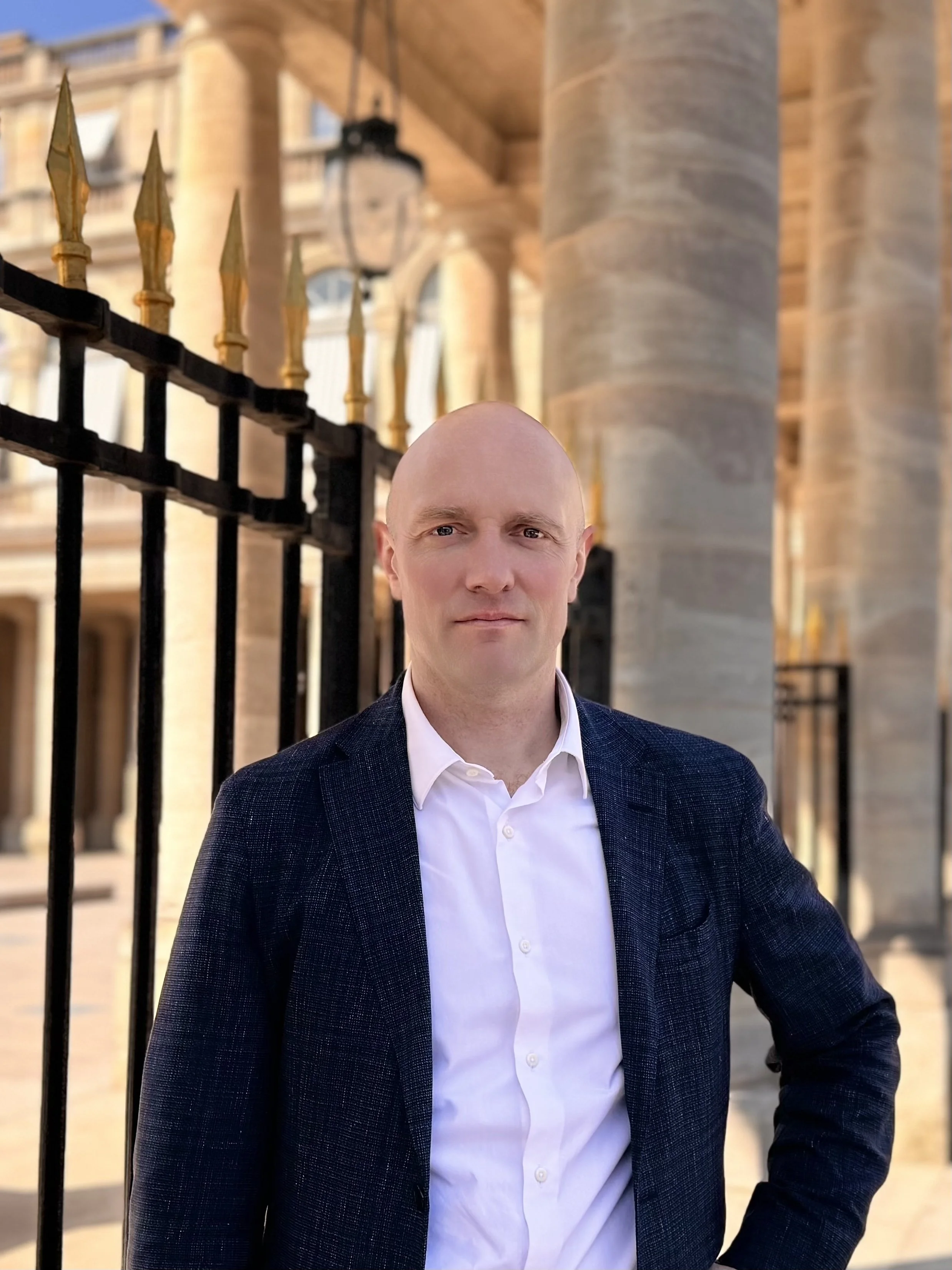 Un homme en veste foncée et chemise blanche pose devant une grille avec des pointes dorées, avec une architecture en pierre en arrière-plan.