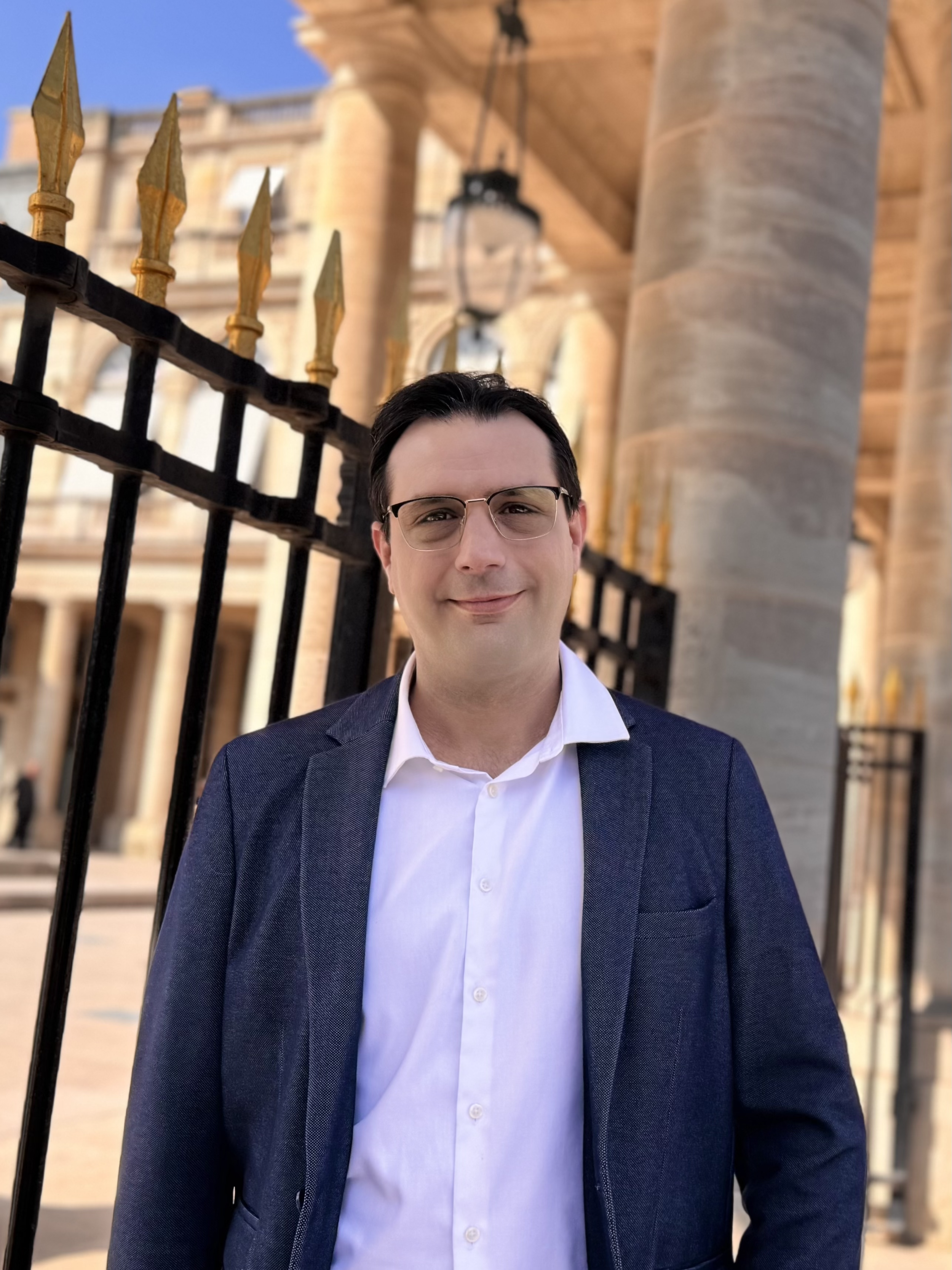 Un homme en costume et lunettes se tient devant une grille noire avec des pointes en or, en arrière-plan un bâtiment en pierre avec colonnes et un lampadaire.
