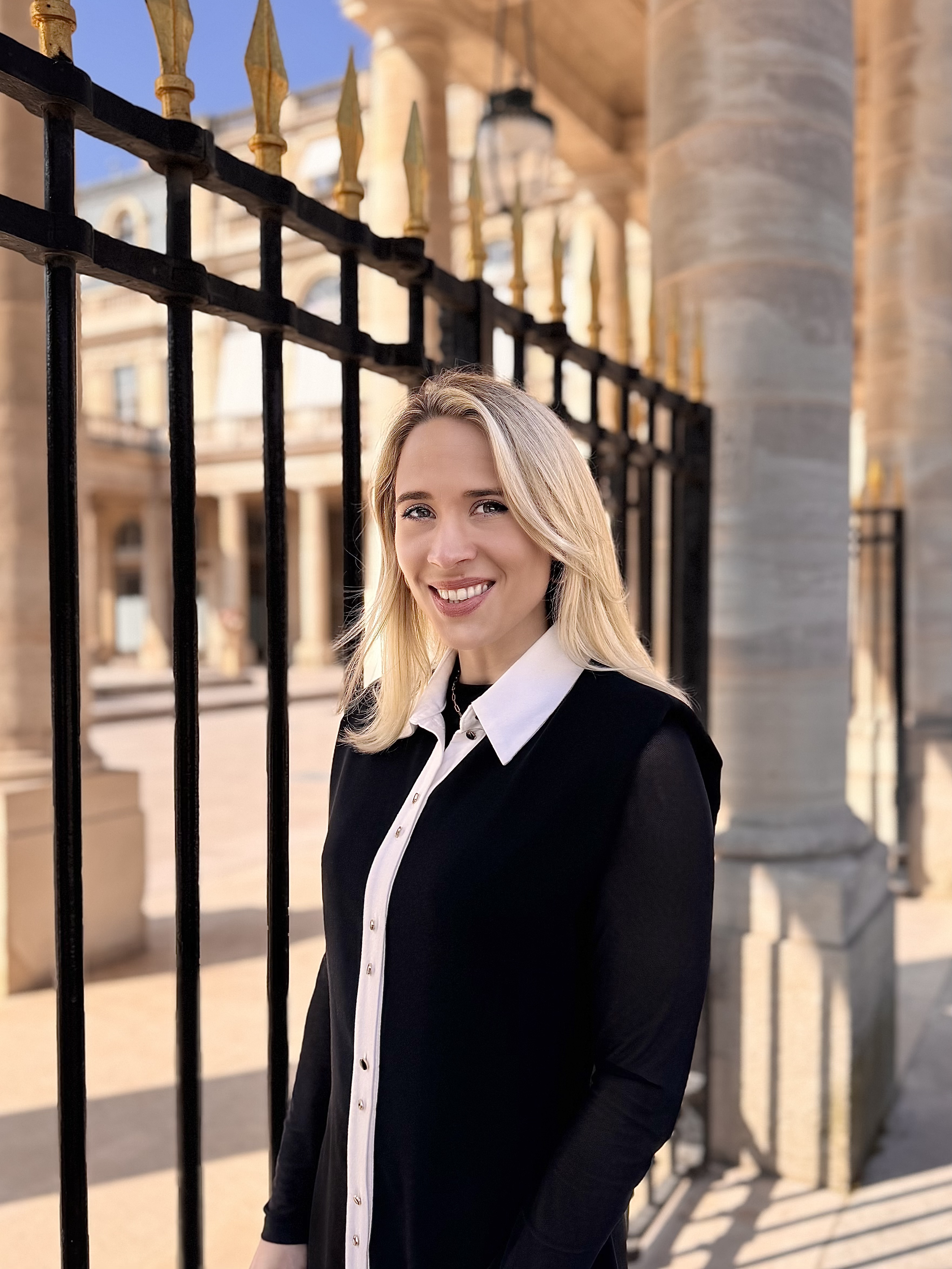 Une jeune femme blonde souriante, portant une chemise noire avec col blanc, posant devant une grille en fer forgé noir avec des détails dorés, en face d'un bâtiment historique en pierre.