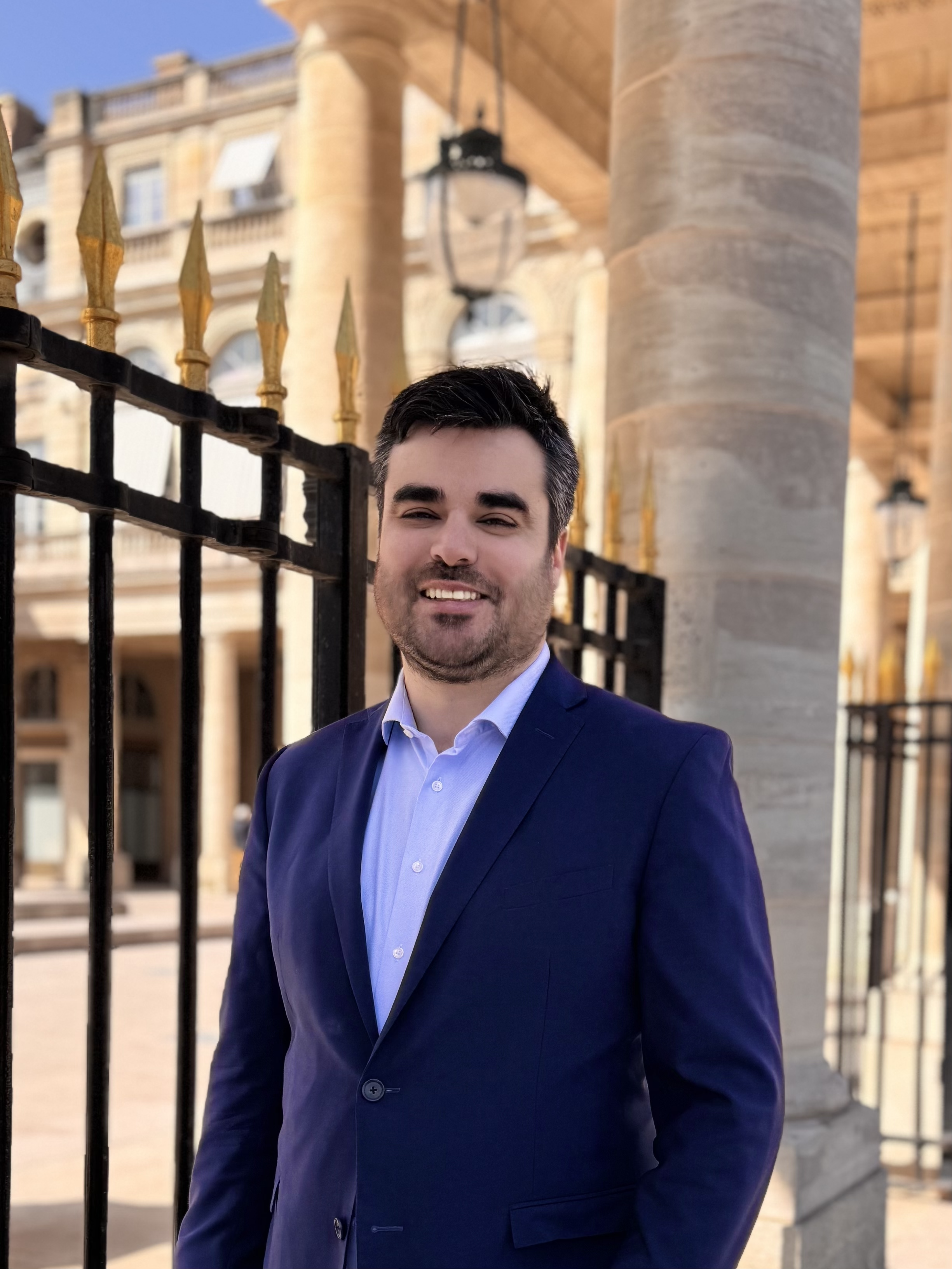 Un homme en costume bleu clair et veste bleu marine, portant une chemise blanche, se tient devant une grille noire avec des pointes dorées, avec un bâtiment en pierre en arrière-plan.