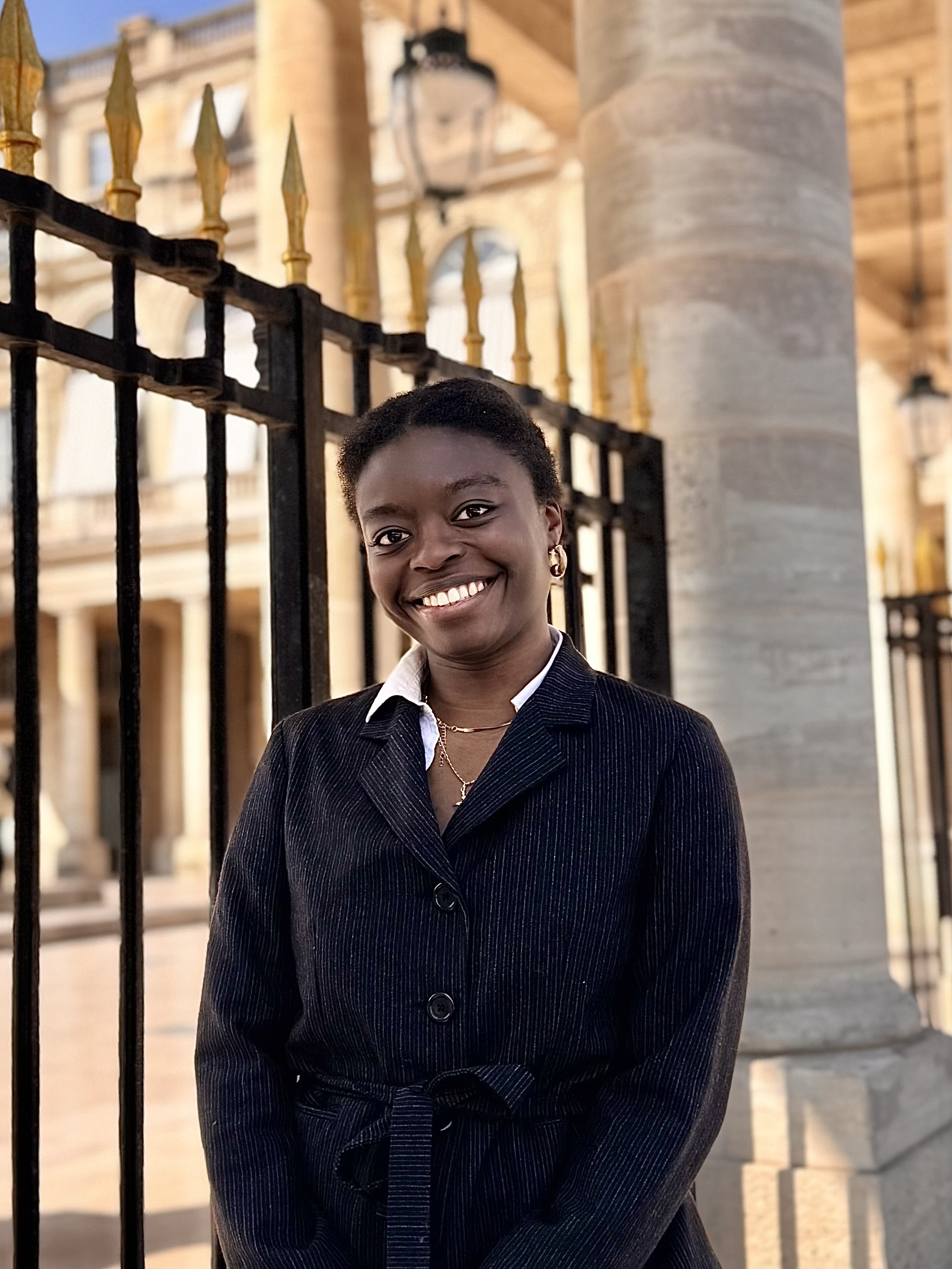 Une femme souriante en costume noir devant une clôture en fer forgé doré et un bâtiment historique avec des lampes extérieures.