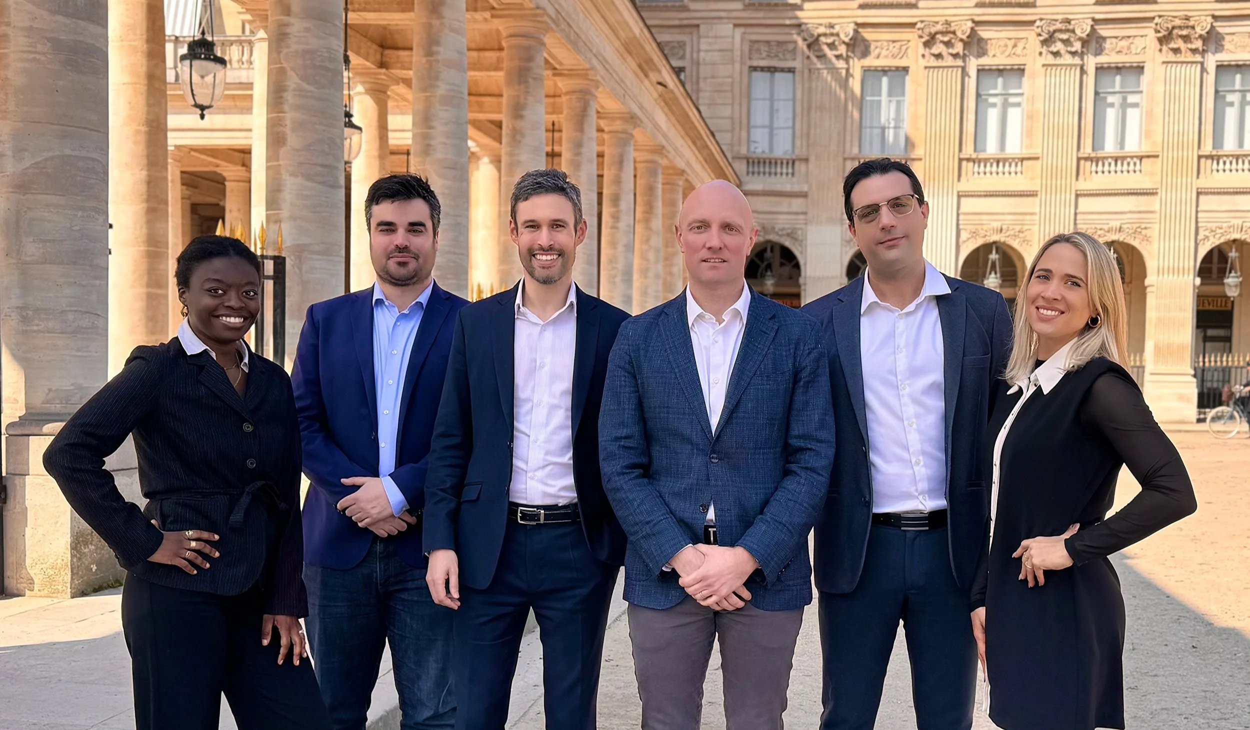 Groupe de six personnes debout devant un bâtiment historique avec des colonnes, tous en tenue professionnelle, souriant, lors d'une journée ensoleillée.
