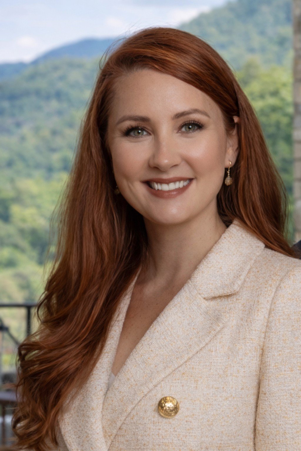 Woman with long red hair smiling outdoors, wearing earrings and a cream-colored blazer with a gold button, with a green, mountainous landscape in the background. She is a realtor from Blue Ridge, GA. The best realtor in Blue Ridge.
