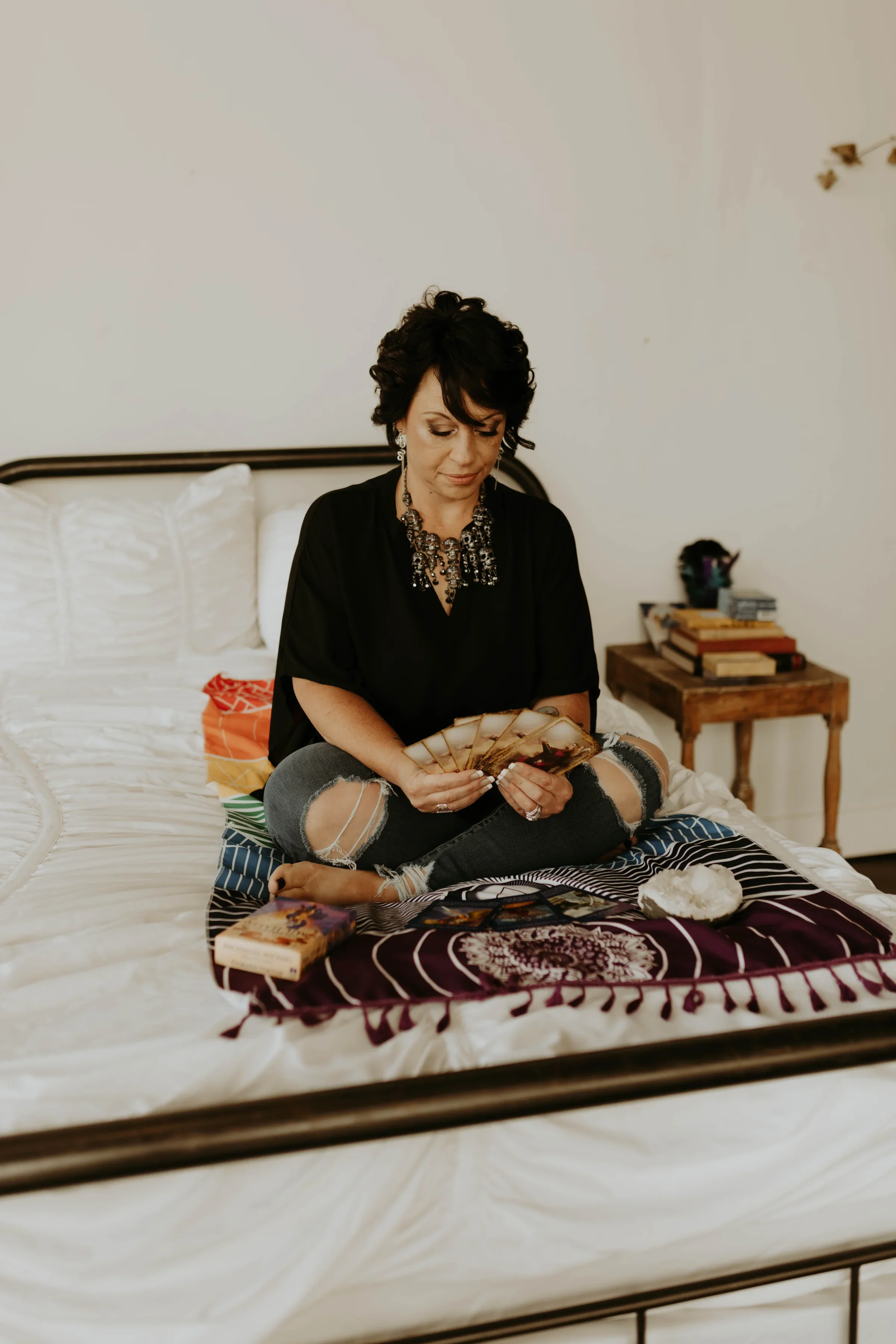 A woman sitting cross-legged on a bed, holding tarot cards, with various books, a crystal, and a tarot deck nearby.