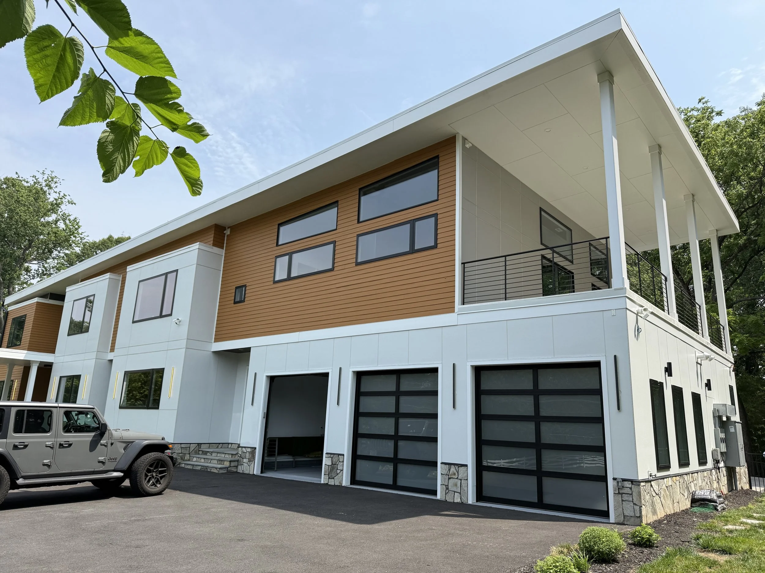 Modern multi-story residential building with large glass windows, a garage, and a third-floor balcony with black railing, surrounded by trees and parking lot.