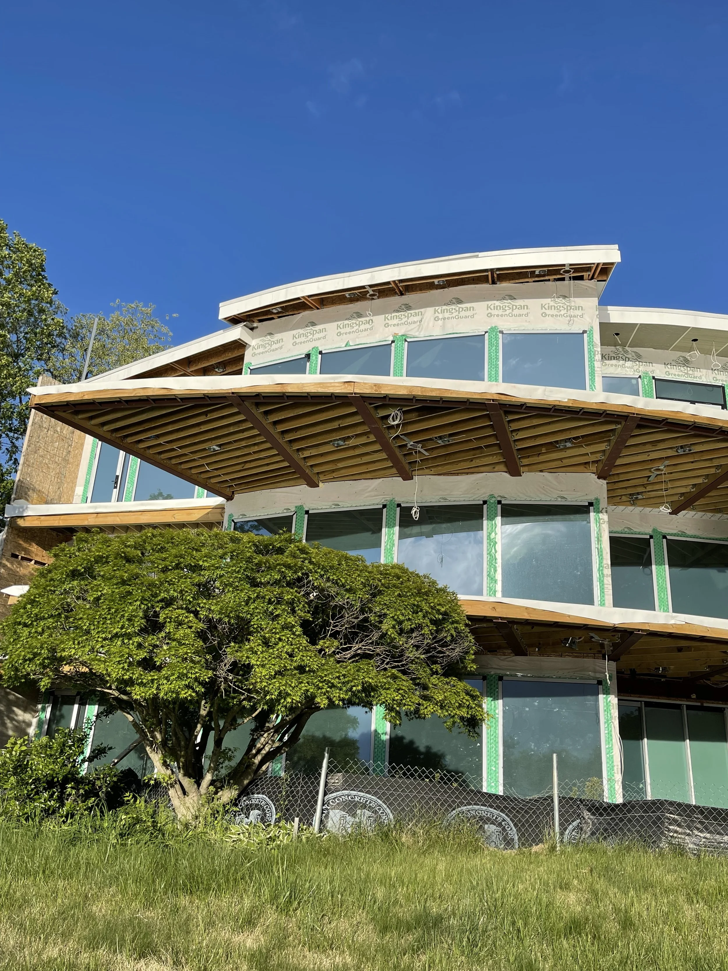 A multi-story building under construction with exposed wooden beams, large glass windows, and green construction wrap, set against a clear blue sky. There is a tree and a grassy area in the foreground with a chain-link fence.