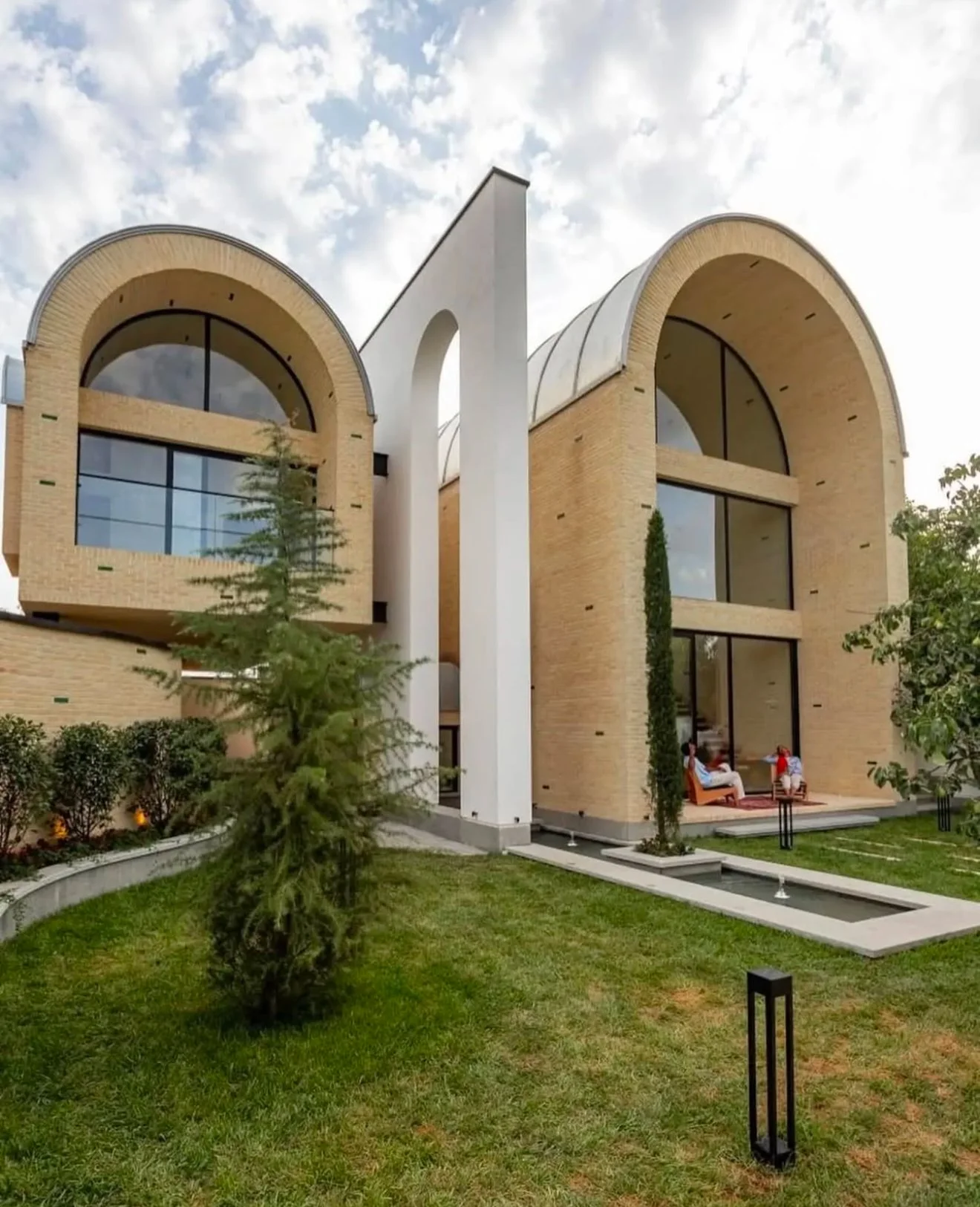 Modern house with large arched windows, brick walls, and a landscaped yard with trees and a pathway, under a partly cloudy sky.