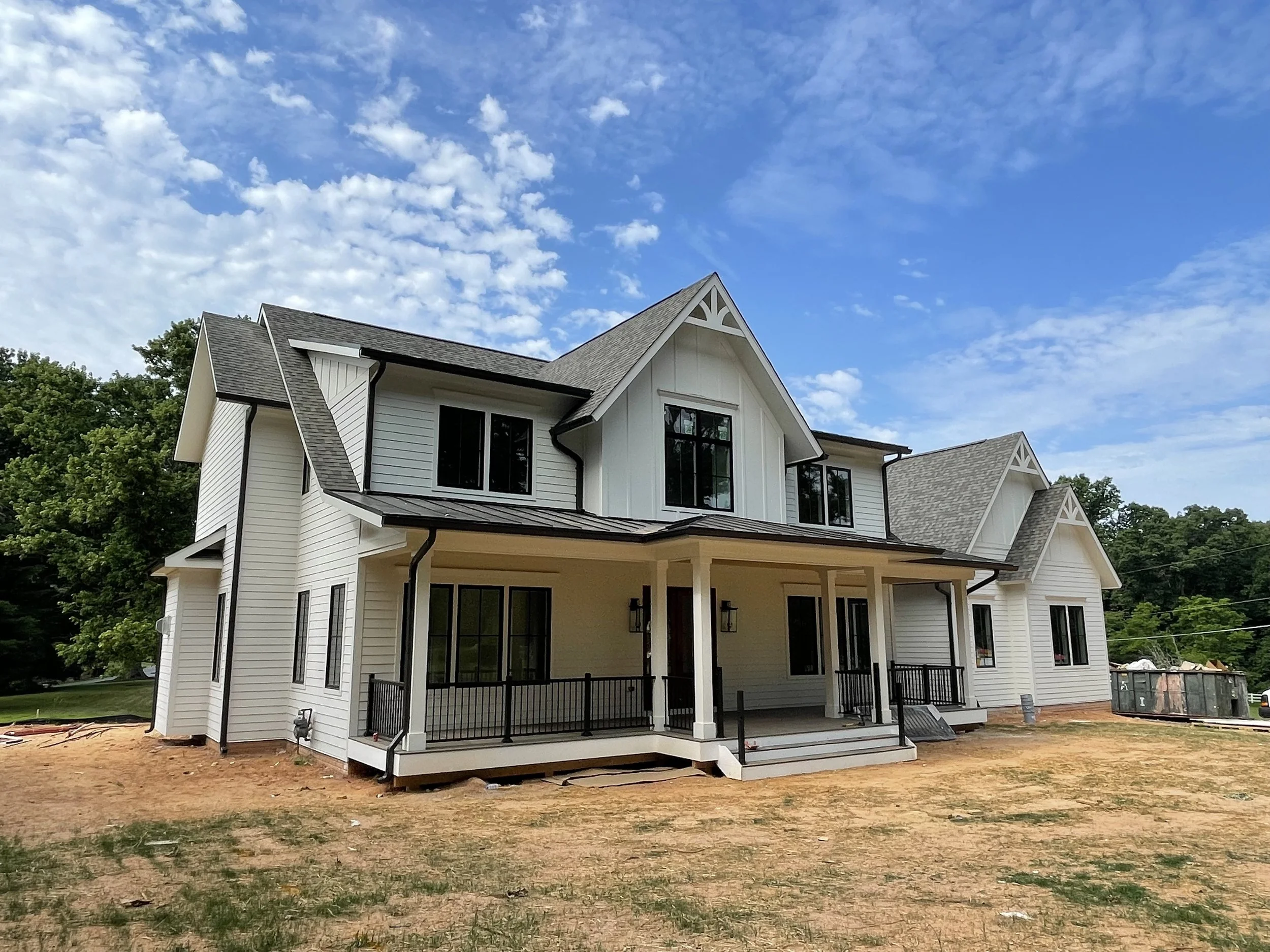 Newly constructed two-story house with white siding, black window frames, and a covered front porch on cleared land with a partly cloudy blue sky overhead.