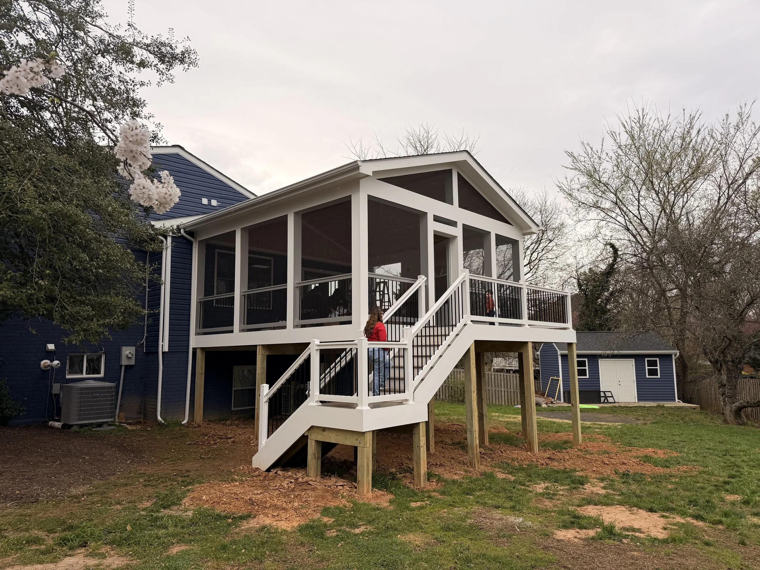 A newly built elevated screened porch with stairs on a backyard, attached to a blue house. A woman in a red jacket is on the stairs. There are trees and a small shed in the background.