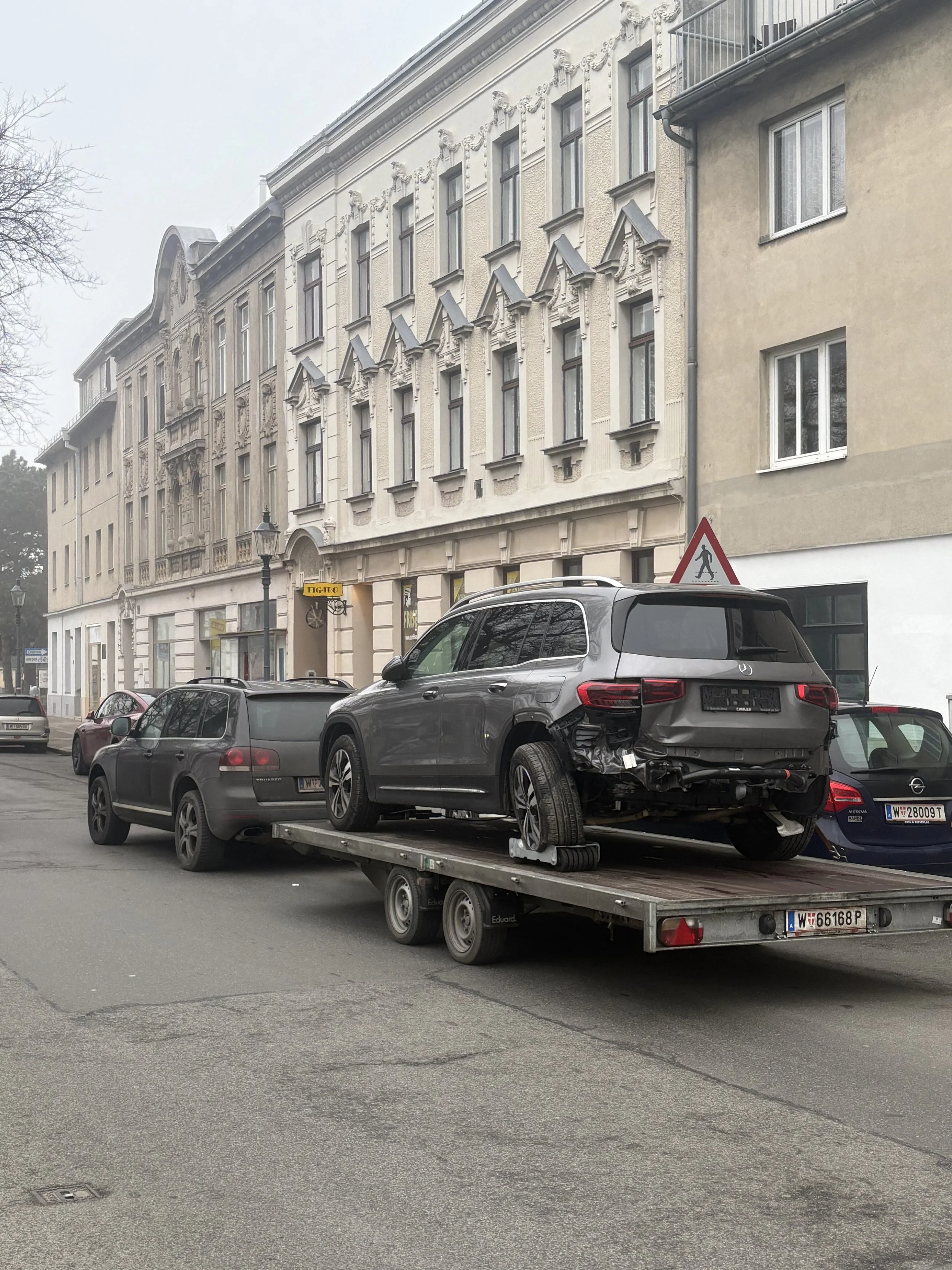 Ein geparkter Lastwagen mit einem schwarzen beschädigten Auto auf einem Stadtstraßenparkplatz, im Hintergrund historische Gebäude mit Verzierungen in Wien, Österreich.