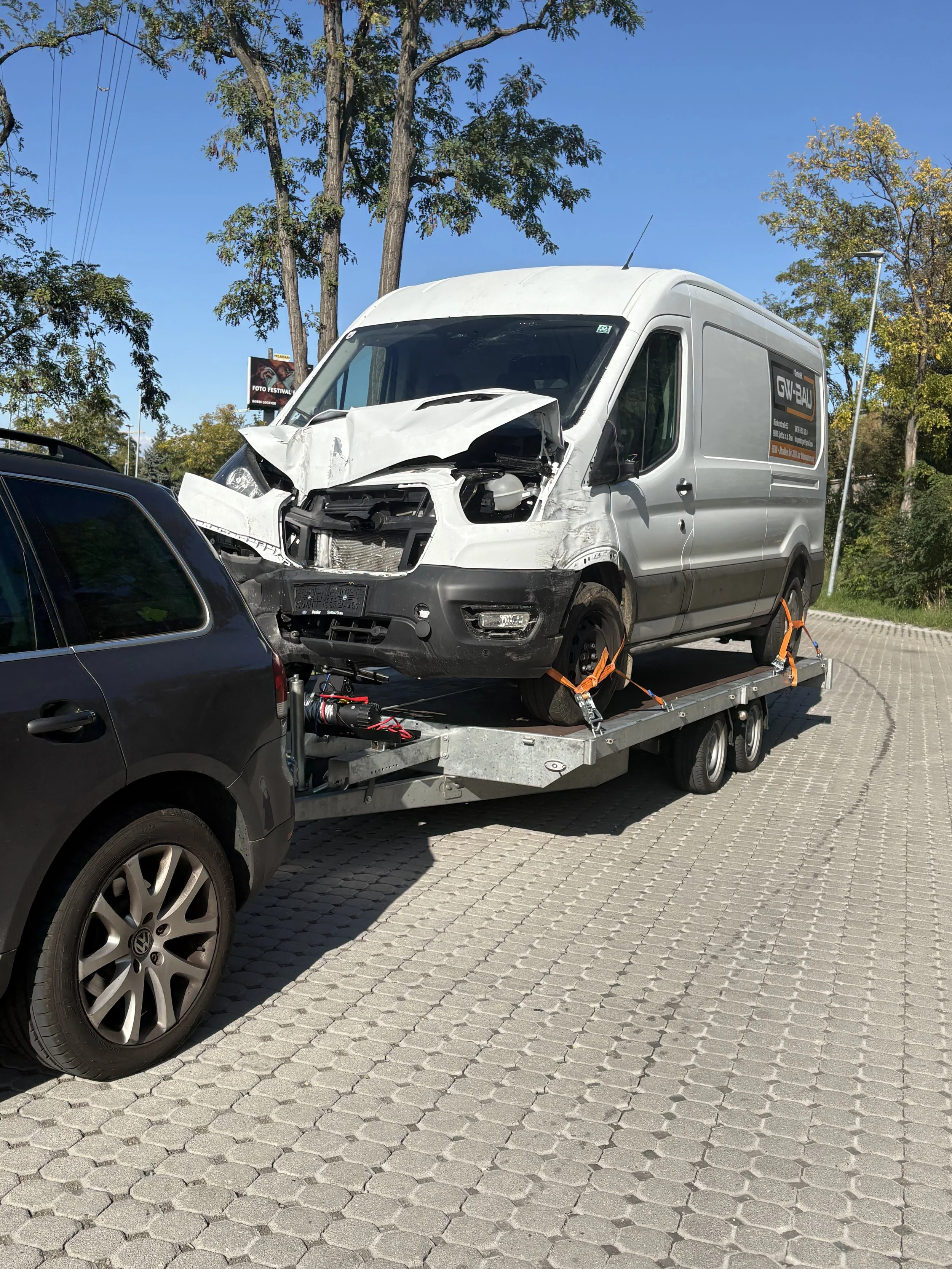 Ein weißer Lieferwagen mit erheblichen Frontschäden, auf einem Abschleppwagen geladen, befindet sich auf einer gepflasterten Straße, umgeben von Bäumen mit grünem Laub und einem blauen Himmel im Hintergrund.